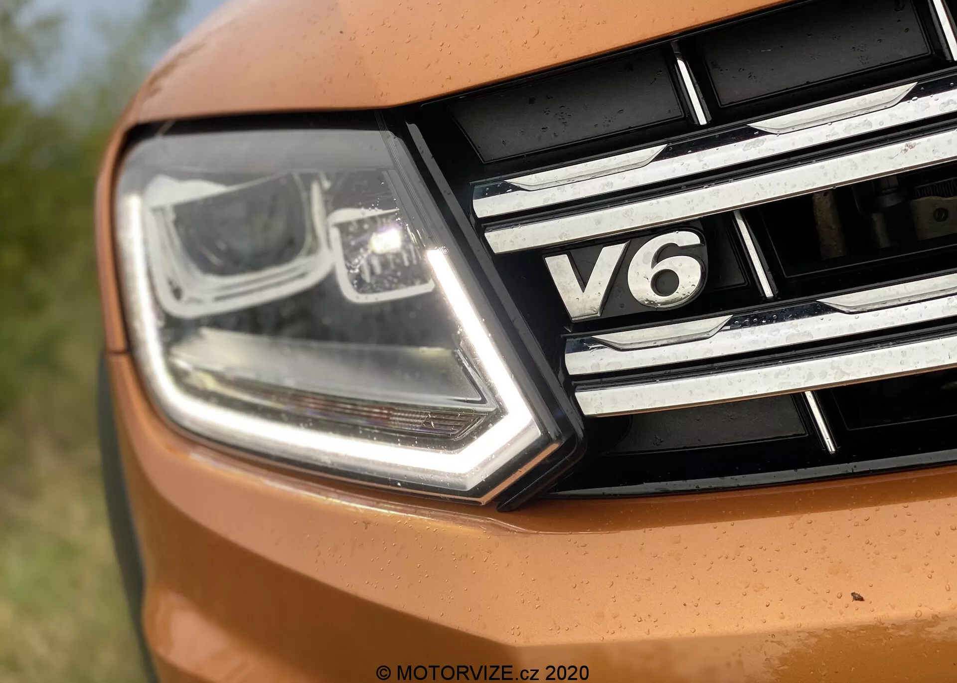 Primo piano della sezione frontale sinistra del VW Nutzfahrzeuge Amarok 2016. L'immagine cattura un faro moderno e angolare con luci diurne a LED e tecnologia a fascio proiettore, insieme a dettagli cromati. La griglia presenta strisce cromate orizzontali e un badge 'V6', indicante il tipo di motore a sei cilindri dell'auto. Parte del cofano appare in una tonalità arancione vibrante, suggerendo un design sportivo.