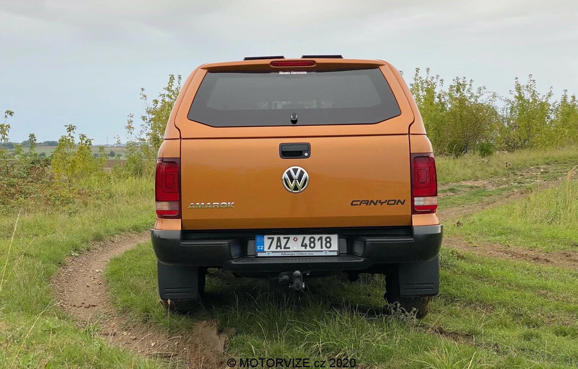 Vista posteriore di un pickup Volkswagen Amarok 2016 arancione metallico parcheggiato all'aperto su un sentiero di terra irregolare con erba ai lati, sotto un cielo coperto. La foto mostra una chiara prospettiva del portellone e delle caratteristiche del design posteriore, incluso il badge Volkswagen, la targhetta Amarok e l'emblema V6. Il veicolo è dotato di paraurti posteriore con gradino integrato, targa centrata e gancio di traino visibile. Sono visibili le luci posteriori orientate verticalmente e la terza luce del freno sulla linea del tetto sopra il parabrezza posteriore, nonché le linee del sbrinatore.