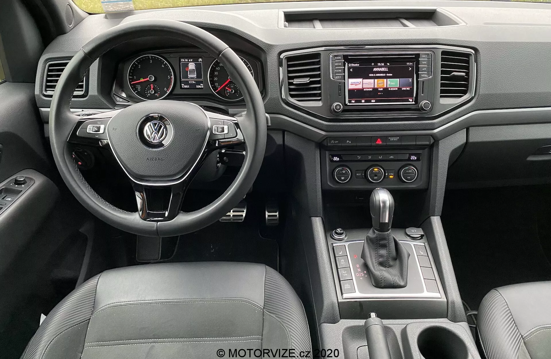 Vista del cruscotto interno del Volkswagen Amarok 2016 dalla prospettiva del conducente. Il volante multifunzione rivestito in pelle con il logo VW è centrale, con quadranti analogici e un display digitale nel quadro strumenti. La console centrale presenta un sistema di infotainment touchscreen, pulsanti per il controllo del clima e una leva del cambio automatico. Dettagli di lusso includono finiture in pelle e cromo, con comandi delle porte visibili.