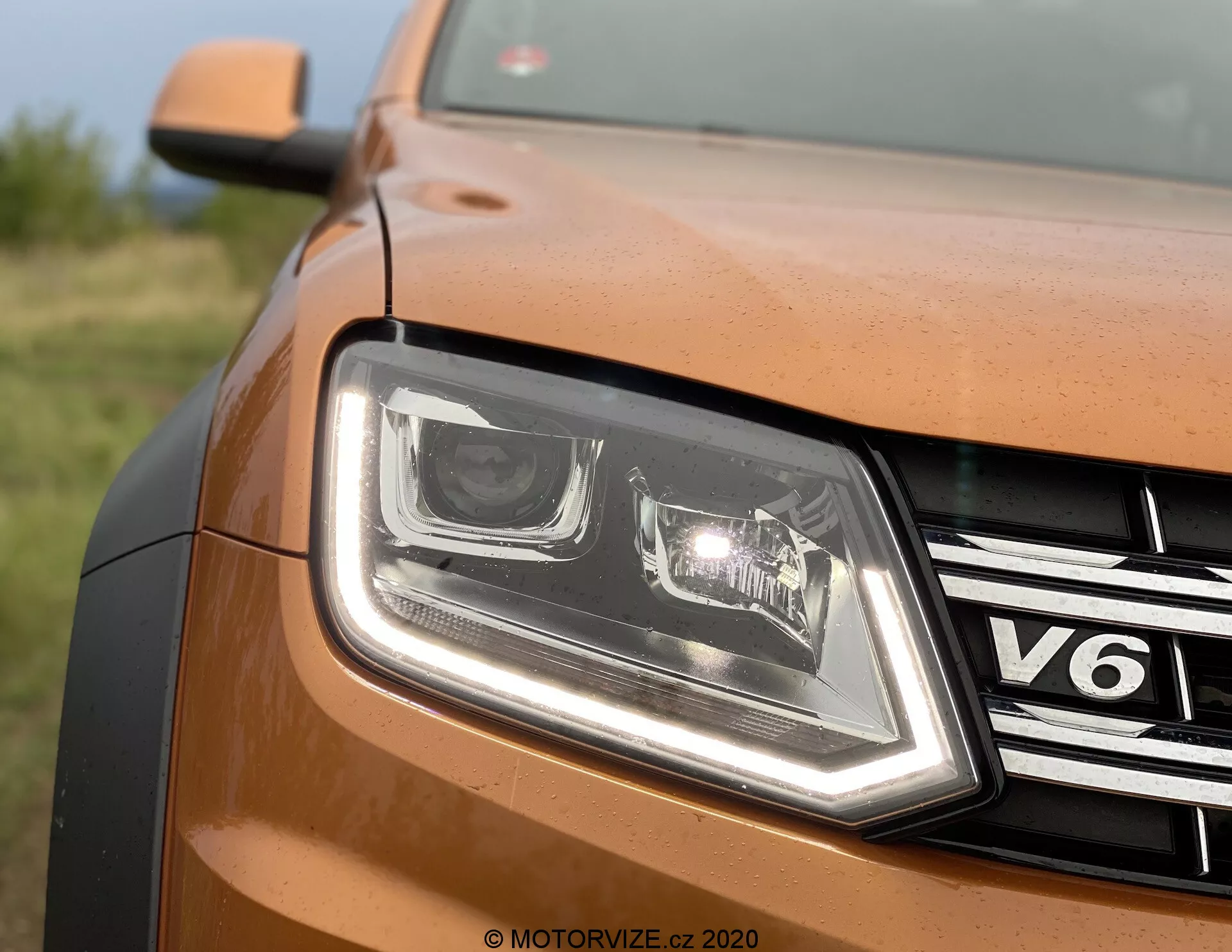 Vista parziale frontale ravvicinata del lato anteriore sinistro di una VW Nutzfahrzeuge Amarok 2016 di colore bronzo o rame. L'immagine mostra il design dei fari a LED con luci diurne distintive, griglia con finitura cromata con badge 'V6', pannello carrozzeria con vernice metallizzata, bordo del cofano sagomato e watermark 'MOTORVIZE.CZ 2020', che implica un'origine di un sito di recensioni ceco.