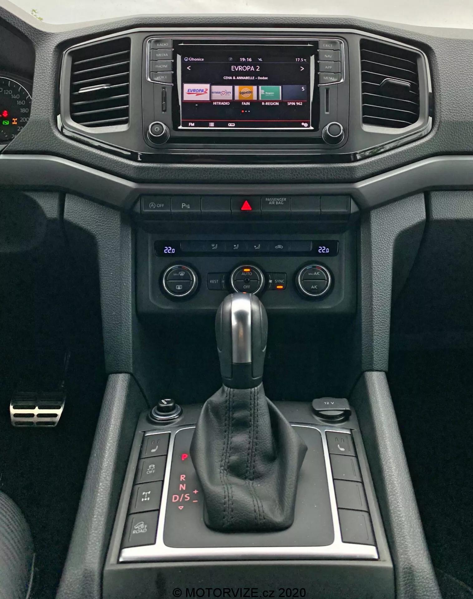 Vista ravvicinata della console centrale moderna del VW Amarok 2016, con in primo piano il sistema di infotainment e il cambio. Il display dell'infotainment suggerisce funzionalità touch, probabilmente mostrando l'output della radio o dei media. Sotto ci sono prese d'aria orizzontali circondate da materiali soft-touch con un design testurizzato. I comandi del clima e gli interruttori sono posizionati sotto le bocchette per regolare la temperatura e il flusso d'aria. Un cambio automatico in modalità 'P' domina il centro, indicando la trasmissione automatica del veicolo con opzioni di cambio manuale. Piccoli scomparti si trovano accanto al cambio, insieme a una manopola girevole, probabilmente per il controllo dell'infotainment o delle modalità di guida. Il design degli interni enfatizza sia la funzionalità che il comfort.