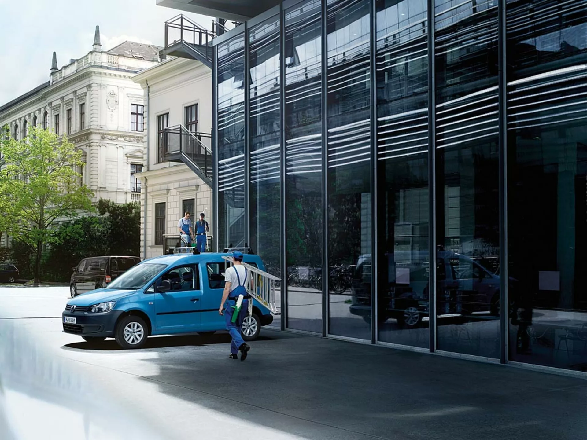 VW Caddy blu del 2010 parcheggiata in strada urbana, mostrata da una vista frontale a tre quarti con le porte aperte e evidenziando la sua griglia frontale, i fari, le ruote e uno scorcio dell'interno su sfondo di architettura moderna e classica
