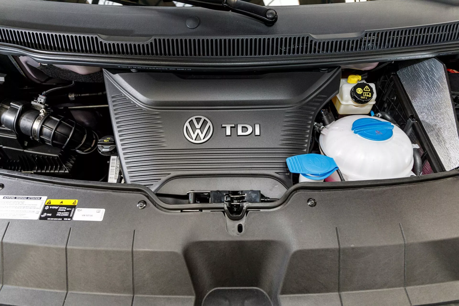 Vista ravvicinata del vano motore diesel della VW Nutzfahrzeuge Transporter 2019 (T6.1 California) mostrando la copertura del motore TDI con logo Volkswagen, serbatoio del refrigerante e batteria dell'auto