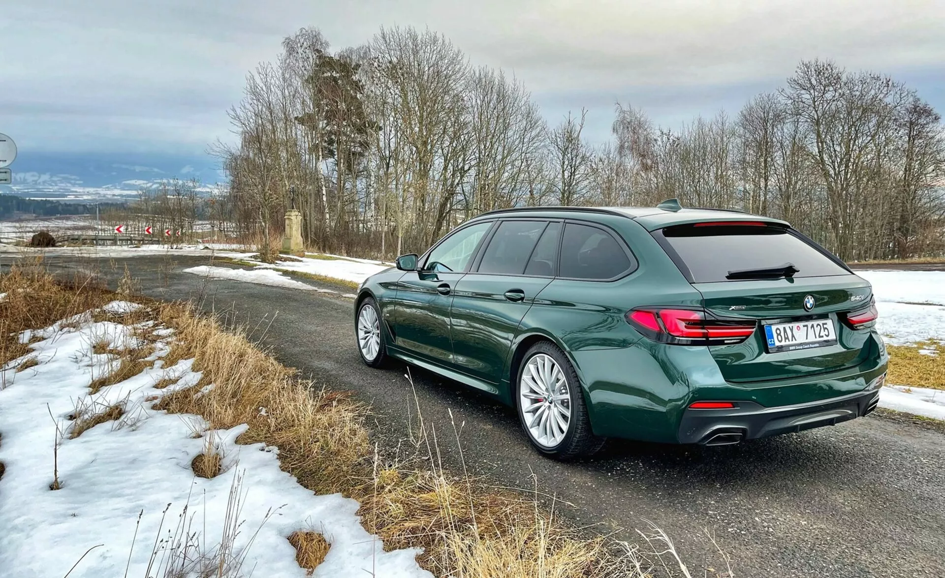Vista posteriore tre quarti di una station wagon BMW che mostra il retro del veicolo con fanali posteriori a forma di L, il profilo laterale sinistro inclusi porte e curvatura del tetto con barre sul tetto, cerchioni in lega con pneumatici stradali, in vernice verde scuro metallizzato, parcheggiata su una strada di campagna con leggera neve, evidenziando elementi di lusso e design moderno.