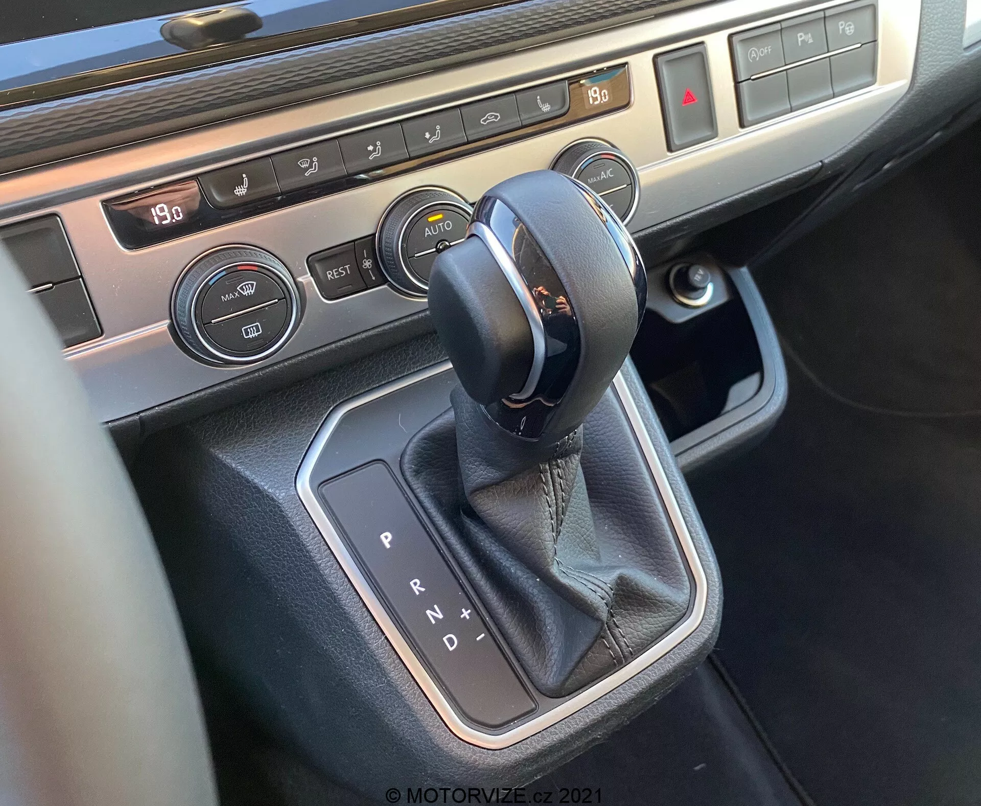 Vista ravvicinata della console centrale del VW Nutzfahrzeuge T6-1 Multivan 2019 focalizzata sull'area della leva del cambio. L'immagine mostra un selettore del cambio automatico con cuciture in pelle, circondato da un rifinitura metallica, pulsanti di controllo del climatizzatore con display della temperatura e comandi per il riscaldamento dei sedili, evidenziando la tecnologia degli interni moderna e i materiali premium del veicolo.