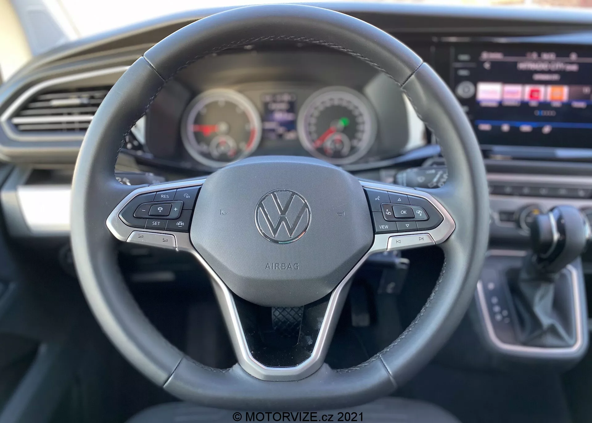 Vista interna di un VW Nutzfahrzeuge Transporter 2019 (T6-1 Multivan) focalizzata sulla prospettiva del conducente. Vista dettagliata del volante con il logo Volkswagen e pulsanti di controllo per varie funzioni come il cruise control e la navigazione del sistema multimediale. Dietro il volante, il quadro strumenti mostra il tachimetro e il contagiri in stile analogico con un display digitale al centro. Console centrale parzialmente visibile con un ampio schermo touch-screen infotainment, insieme a un design elegante del cruscotto con presa d'aria orizzontale. La leva del cambio a destra indica una trasmissione automatica. L'immagine evidenzia le funzioni di usabilità con una profondità di campo ridotta focalizzandosi principalmente sul volante.
