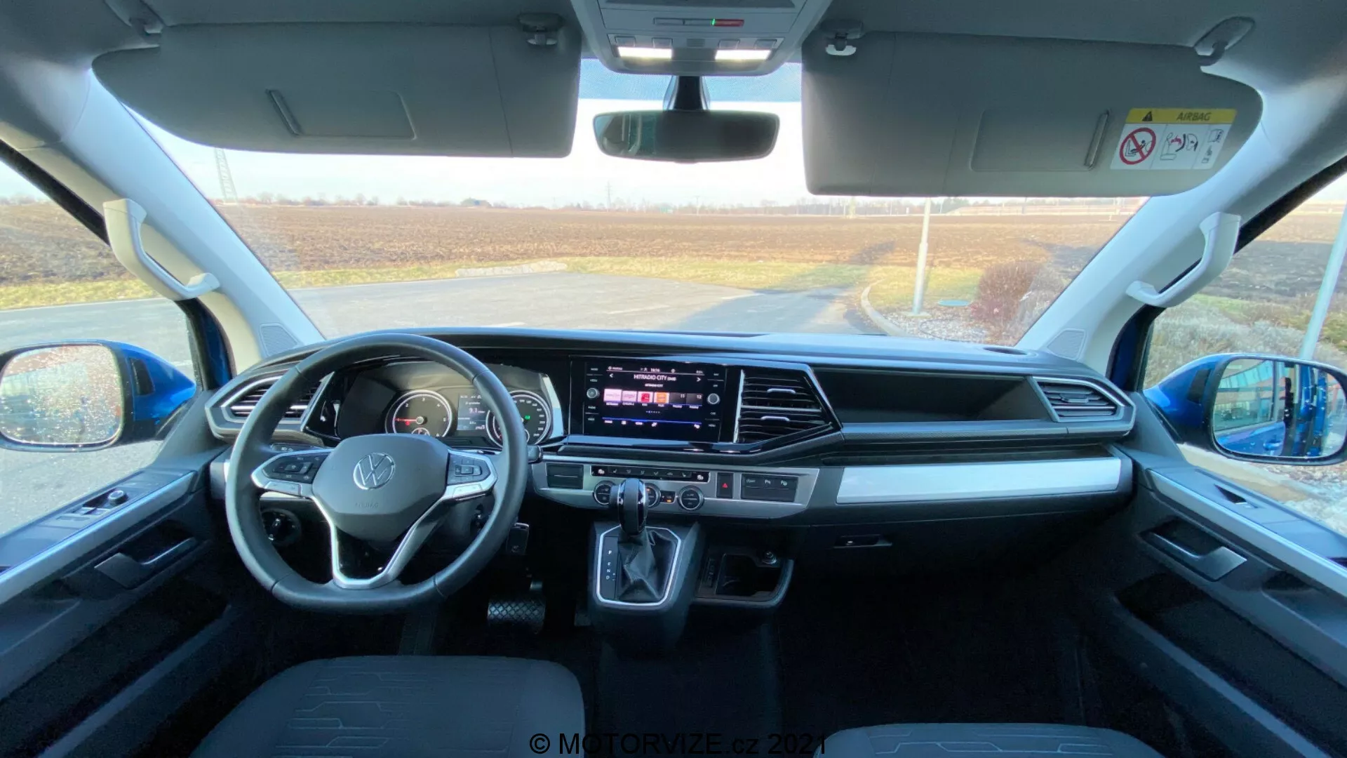 Interni di un VW Nutzfahrzeuge Transporter 2019 (T6.1 Multivan), ripresi dall'area dei sedili posteriori guardando verso il cruscotto anteriore. La foto è scattata durante il giorno con luce naturale che entra attraverso il parabrezza anteriore, fornendo una vista dettagliata dello spazio abitativo, i sedili anteriori, il volante con comandi montati, lo schermo touchscreen centrale dell'infotainment, i pulsanti e le manopole HVAC, la leva del cambio automatico, gli spazi di stoccaggio, i porti di connettività, i controlli delle porte e degli specchi, le caratteristiche di sicurezza e il design confortevole dei sedili.