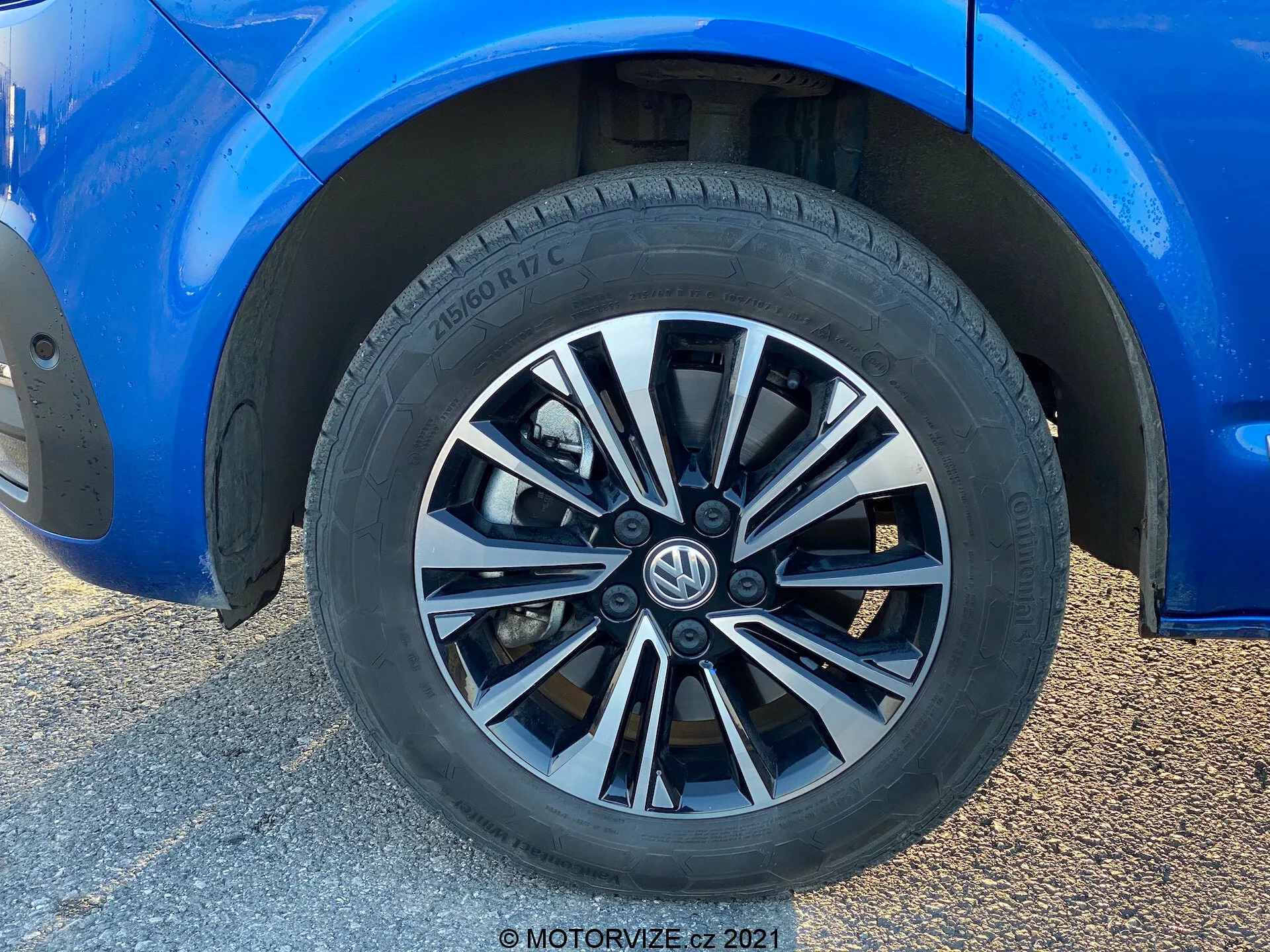 Vista ravvicinata di una ruota del Volkswagen T6-1 Multivan 2019. Questa immagine a pieno fotogramma presenta un design della ruota a più raggi con finiture alternanti in argento e scuro. Il pneumatico, di marca Hankook, mostra un disegno del battistrada per prestazioni su strada e specifiche parziali del pneumatico. La carrozzeria blu circostante, in condizioni impeccabili, riflette una lucentezza trovata. Si denota un'altezza da terra moderata e pinze e rotori dei freni standard parzialmente visibili. La luce naturale esalta la composizione focalizzata sulla ruota, con lo sfondo sfocato a causa di una ripresa ravvicinata a basso angolo.