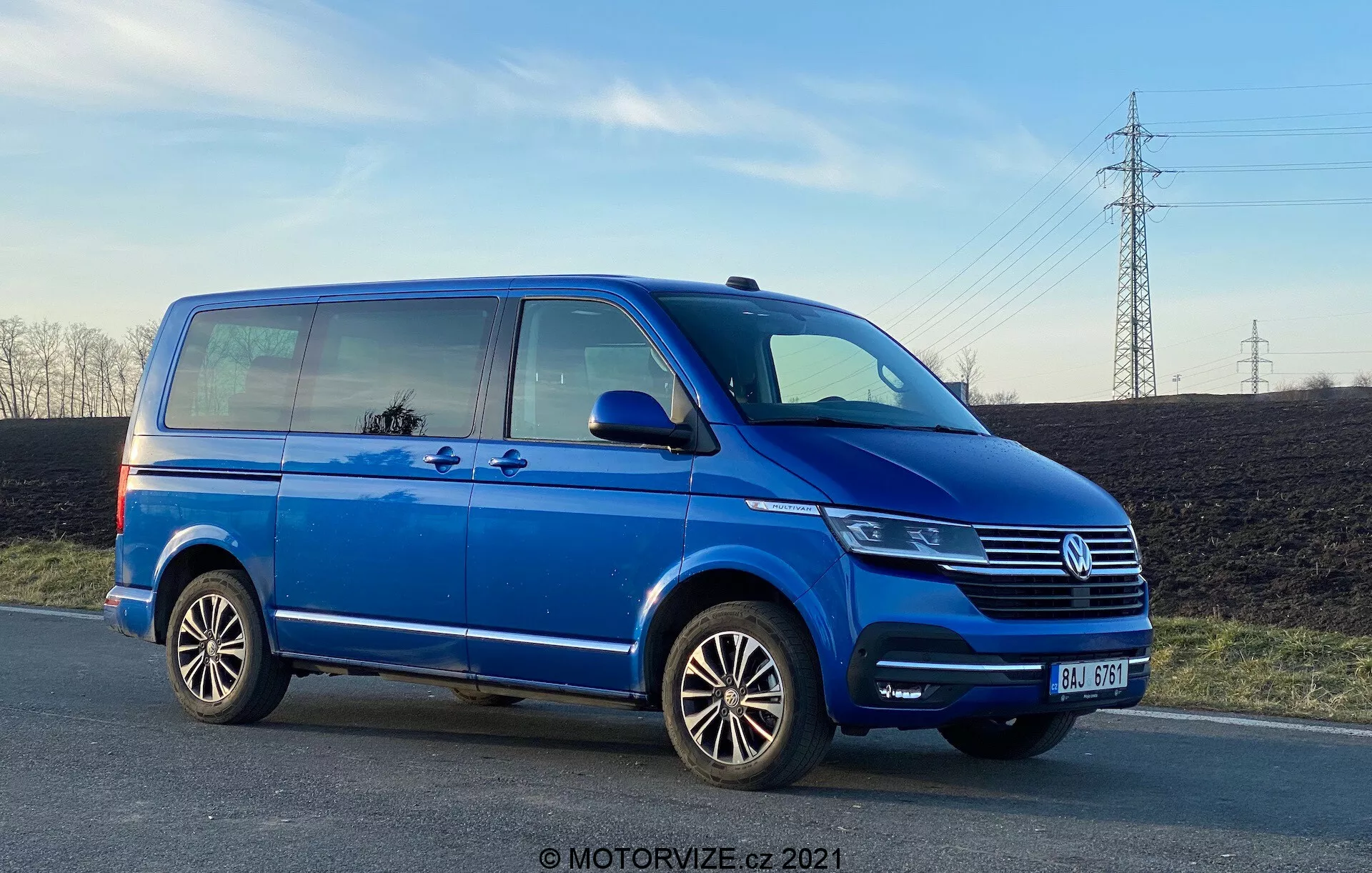 Un furgone Volkswagen Transporter T6 blu, che mostra il profilo laterale da un angolo basso, enfatizzando la lunghezza e l'altezza. L'immagine presenta dettagli come la porta scorrevole del passeggero, la porta anteriore del passeggero, la ruota posteriore lato passeggero, cerchi in lega multirazze, e vedute parziali del faro, della griglia Volkswagen con emblema e del paraurti anteriore. La foto, scattata nel tardo pomeriggio, mostra l'altezza da terra e riflette il cielo e i dintorni nelle finestre, con un cielo limpido e linee elettriche sullo sfondo.
