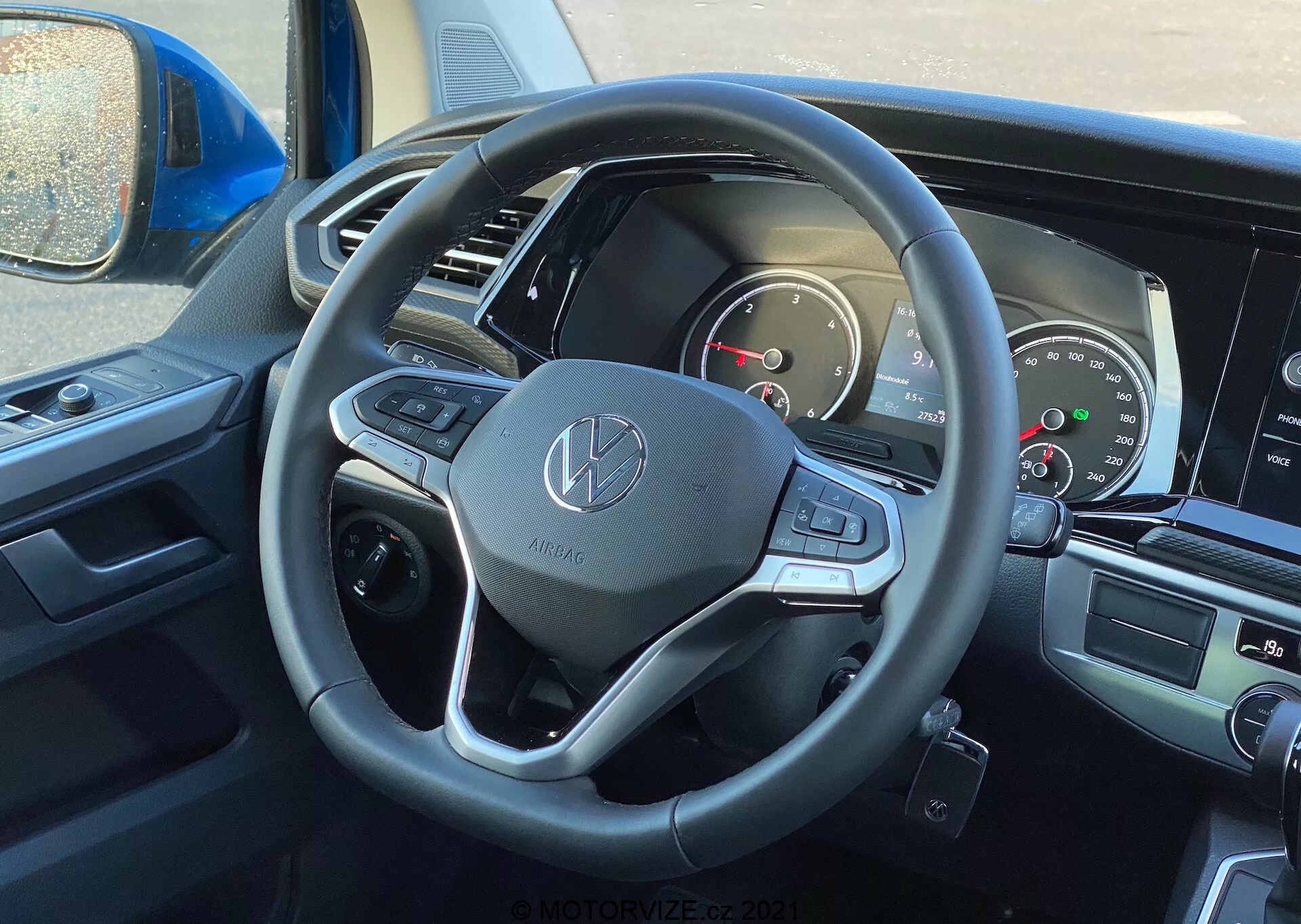 Vista interna del VW Nutzfahrzeuge Transporter 2019 (T6-1 Multivan) dalla prospettiva del guidatore, con attenzione al volante e al cruscotto. Il volante mostra l'emblema Volkswagen ed è rivestito in pelle, con pulsanti multifunzionali e paddle di cambio. Il quadro strumenti parzialmente visibile include quadranti analogici e uno schermo digitale. La schermata centrale dell'infotainment e i controlli del climatizzatore sono leggermente visibili, indicando funzionalità moderne di multimedialità e comfort.