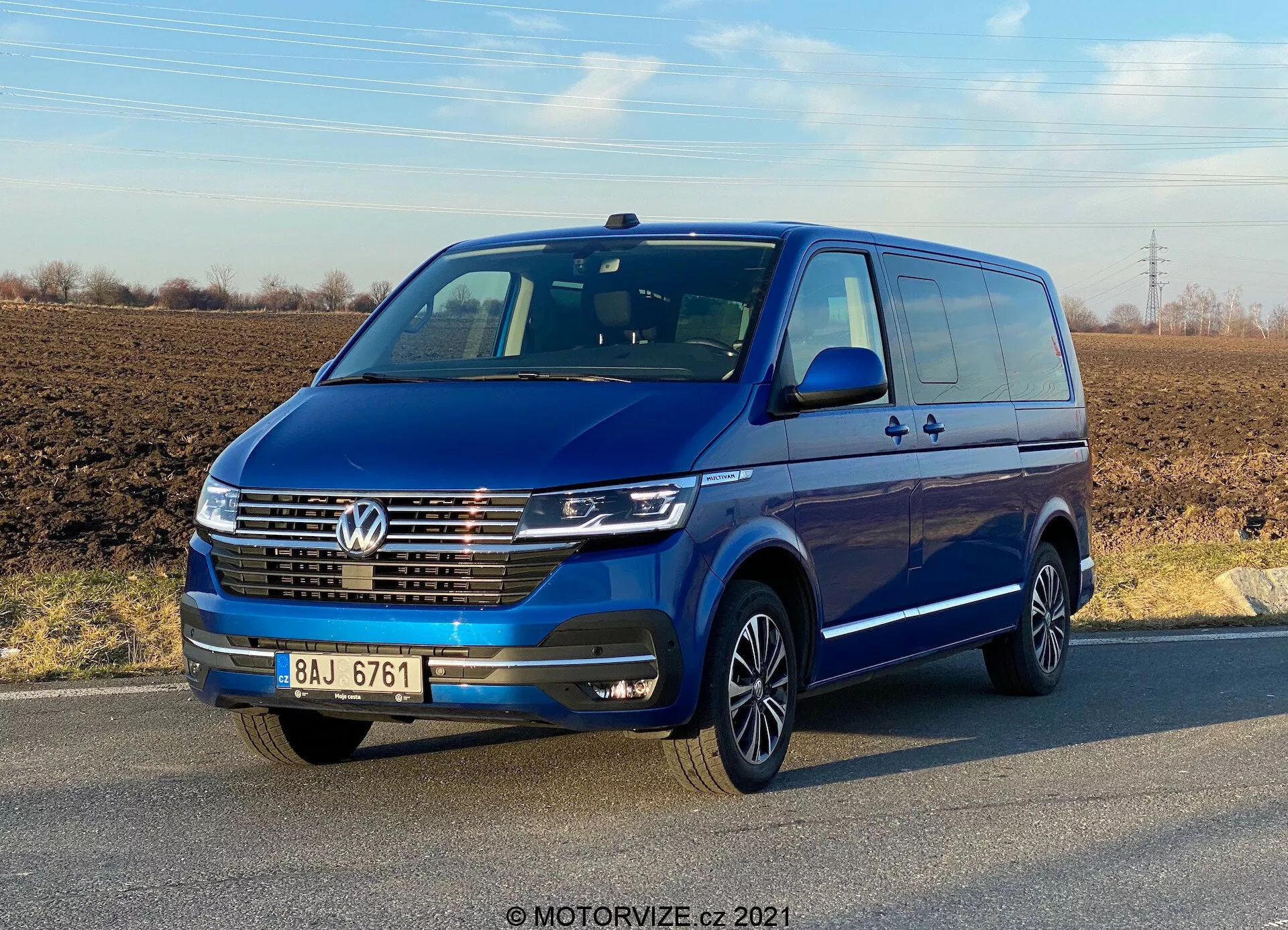 Vista tre quarti anteriore di un Volkswagen Transporter T6.1 Multivan blu 2019 sotto un cielo limpido. Il veicolo è dotato di emblema Volkswagen sulla griglia, luci diurne a LED, rifiniture cromate sulla griglia anteriore e cerchi in lega multiraggio. Specchietti e paraurti in tinta carrozzeria suggeriscono un modello ad alta specifica. Sono visibili le portiere e le maniglie delle portiere, inclusa una porta scorrevole posteriore, che indica la capacità per più passeggeri o carico.