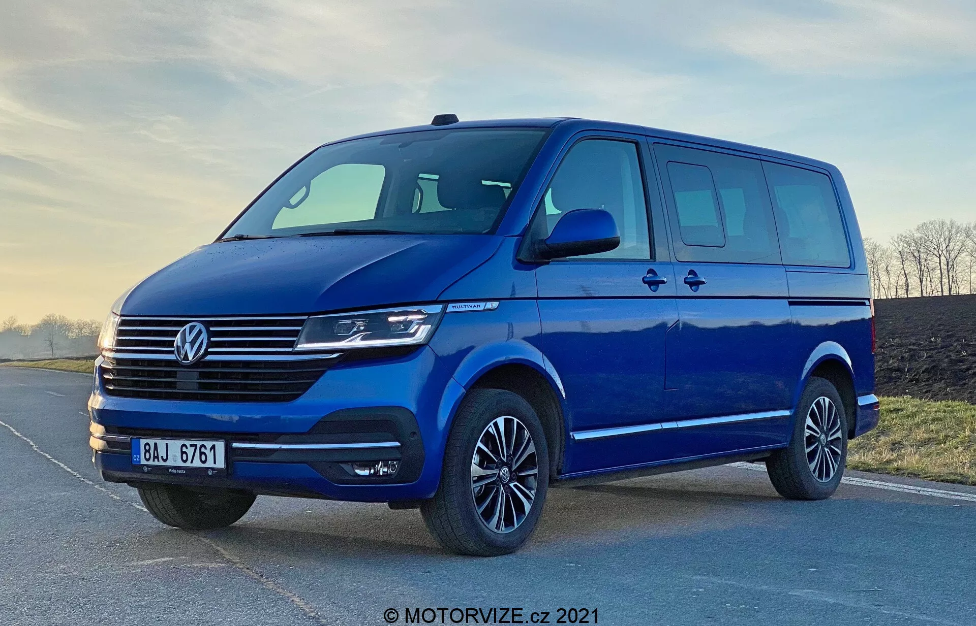 Immagine di un furgone blu Volkswagen Transporter T6.1 Multivan del 2019, ripresa da un'angolazione bassa per accentuarne l'altezza e la dimensione. La vista frontale tre quarti mostra il design frontale e laterale del veicolo, inclusa la distintiva griglia Volkswagen con linee orizzontali, il logo centrale, fari moderni che probabilmente includono tecnologia LED o Xenon, e un paraurti anteriore elegante con un'ampia presa d'aria. Gli specchi laterali aerodinamici con indicatori di direzione integrati, l'ampio spazio finestrato con una porta scorrevole laterale, cerchi in lega con pneumatici a profilo ribassato e un'illuminazione soffusa evidenziano la praticità e l'appeal estetico del furgone. La filigrana 'MOTORVIZE.cz 2021' suggerisce che l'immagine sia del 2021.