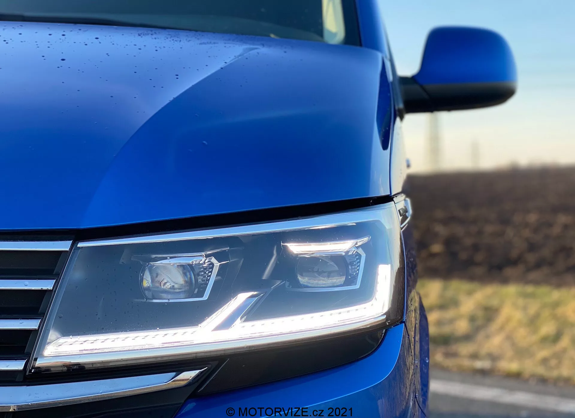 Primo piano angolato basso della sezione anteriore sinistra del VW Nutzfahrzeuge Transporter 2019 (T6.1 Multivan) in blu, con fari LED moderni e angolari con luci diurne, parte della griglia cromata e uno scorcio del contorno del cofano e dello specchietto laterale. La foto, scattata con una profondità di campo ridotta, enfatizza il faro e la carrozzeria circostante, mettendo in evidenza la tecnologia avanzata di illuminazione, il design contemporaneo e la qualità della verniciatura, completato da gocce di pioggia che indicano un rivestimento esterno dettagliato.