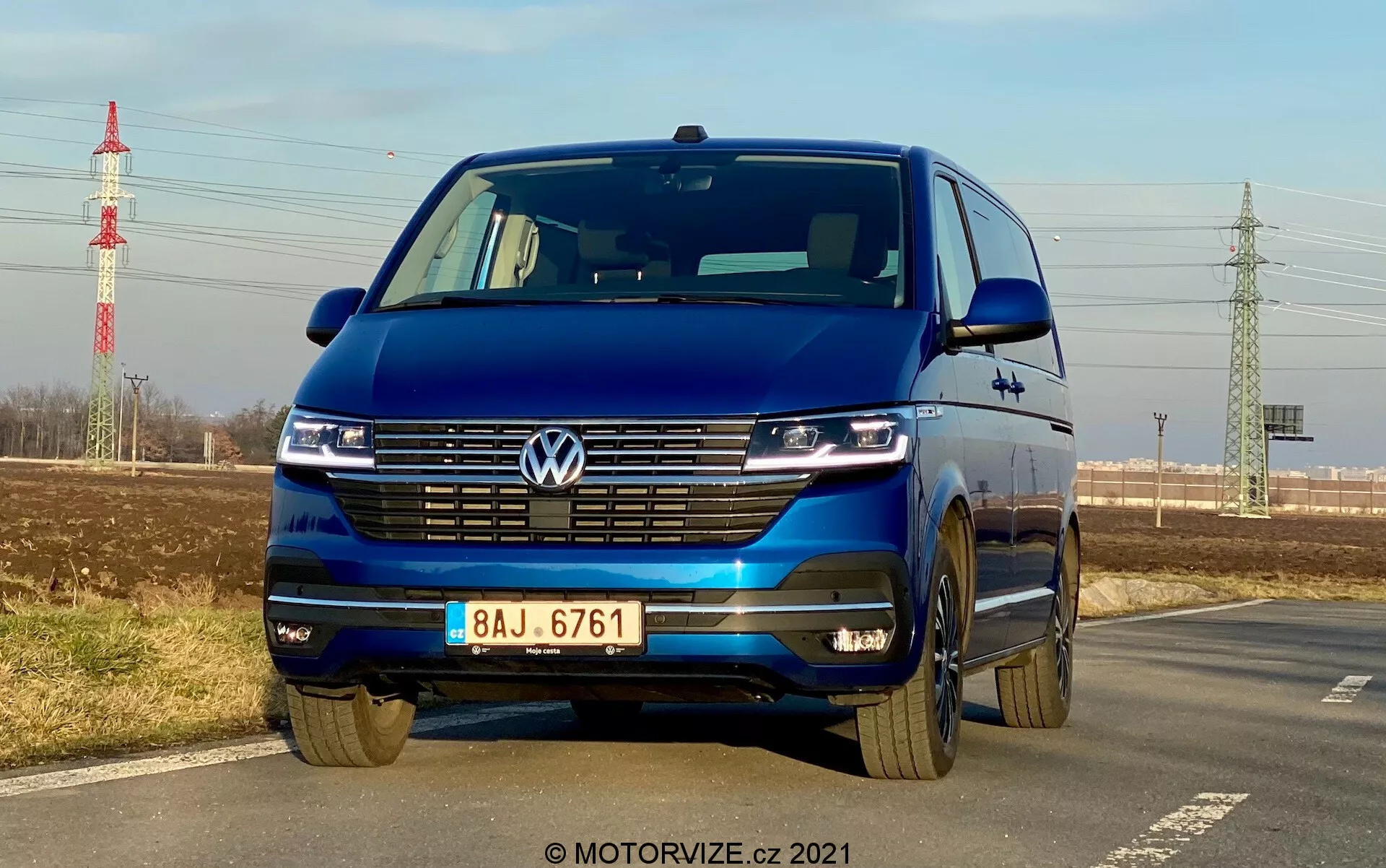 Volkswagen T6-1 Multivan 2019 blu catturato da un angolo frontale basso che evidenzia la griglia, i fari LED con luci diurne, il logo Volkswagen, la griglia larga, il paraurti bicolore e i cerchi in lega; parcheggiato sul lato di una strada con cielo limpido, vegetazione lontana, traliccio della corrente e infrastrutture sullo sfondo.
