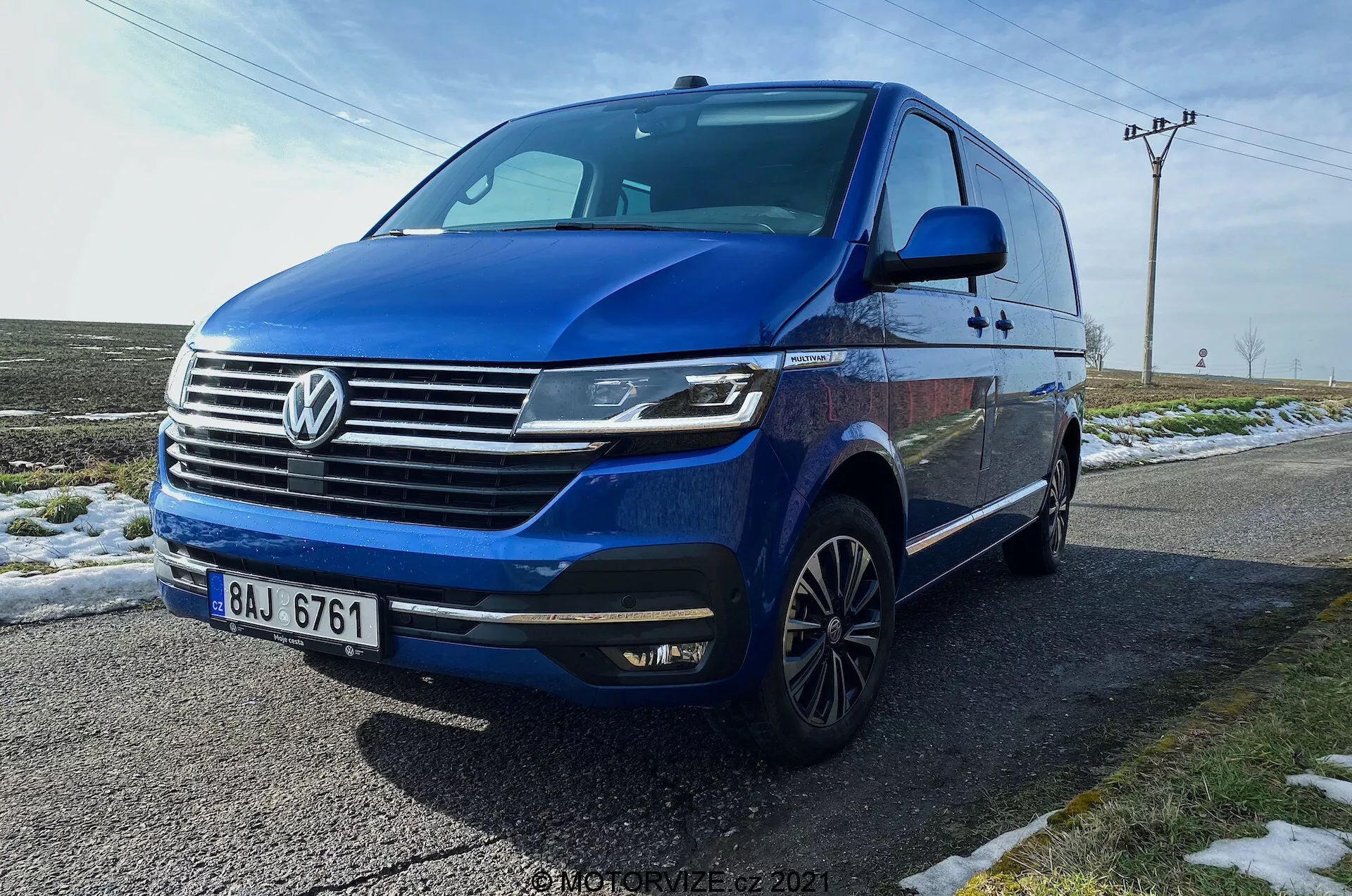Volkswagen T6.1 Multivan Transporter 2019 blu catturato da un angolo frontale a tre quarti basso, mostrando la parte anteriore dell'auto, griglia con logo Volkswagen prominente, barre cromate orizzontali, fari LED angolari con luci diurne, paraurti anteriore con luci antinebbia integrate e prese d'aria, linea dinamica che si estende sui lati, ruote in lega multirazze, frecce integrate negli specchietti laterali, finestre oscurate. Il veicolo è parcheggiato su una strada bagnata accanto a erba innevata, con uno sfondo rurale che include pali della luce e cieli aperti.