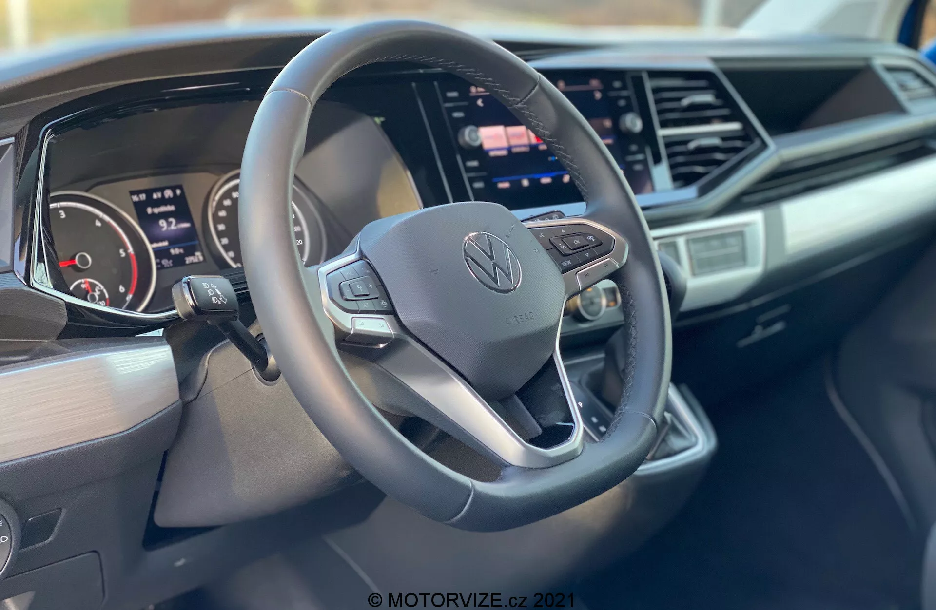 Scatto degli interni di un Volkswagen Nutzfahrzeuge Transporter 2019 (T6-1 Multivan) concentrato sul volante e sul cruscotto. Il primo piano, ripreso dal lato passeggero, mette in evidenza un volante a tre razze con fondo piatto con pulsanti di controllo e logo Volkswagen al centro, un quadro strumenti digitale, un ampio schermo touchscreen di infotainment, un design elegante del cruscotto con accenti angolari e marchio sul lato passeggero del cruscotto.