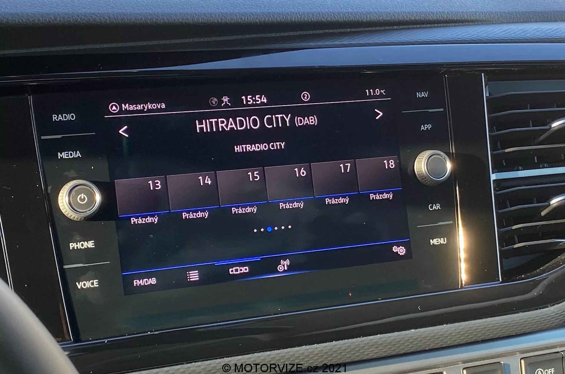 Primo piano frontale dello schermo del sistema di infotainment nel VW-Nutzfahrzeuge Transporter 2019 (T6-1 Multivan). L'immagine presenta in primo piano la stazione radio digitale 'HITRADIO CITY' selezionata, con varie opzioni touch e pulsanti funzione come 'PHONE', 'VOICE' e 'MENU'. Lo schermo mostra l'ora '15:54' e la temperatura esterna '11.0°C', con icone per i profili utente, impostazioni del veicolo e l'intensità del segnale. Lo schermo è integrato nella parte centrale del cruscotto, circondato da pulsanti tattili e manopole di controllo.