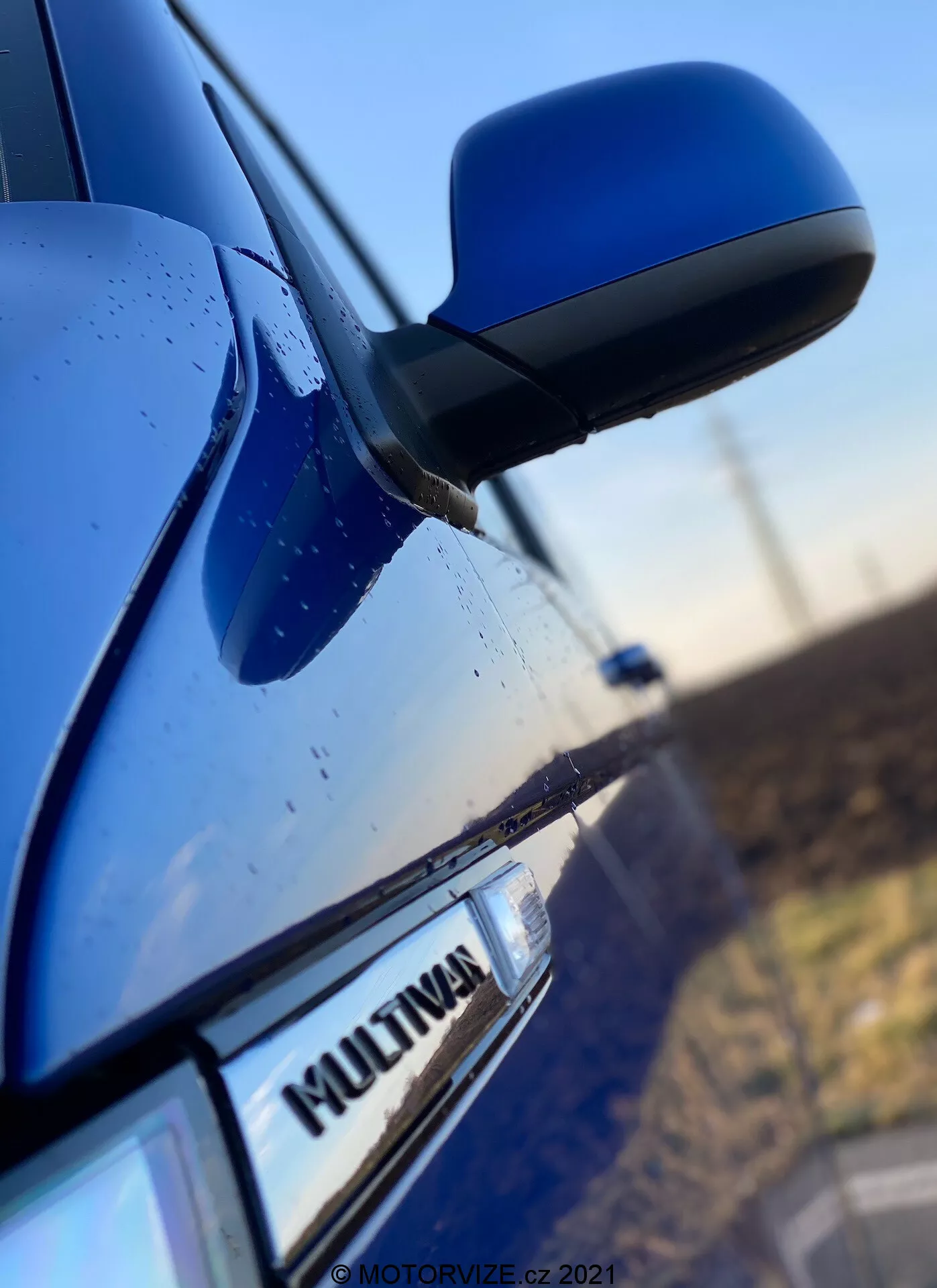 Immagine ravvicinata del lato di un VW Nutzfahrzeuge Transporter 2019 blu (T6-1-Multivan) che mostra parte della porta del passeggero anteriore, lo specchietto laterale e una parte della porta posteriore. L'attenzione è concentrata sul badge 'MULTIVAN' dell'auto, con lo specchietto laterale aerodinamico leggermente sfocato. La carcassa dello specchietto corrisponde al colore della carrozzeria e le gocce d'acqua sulla superficie suggeriscono caratteristiche impermeabili o pioggia recente.