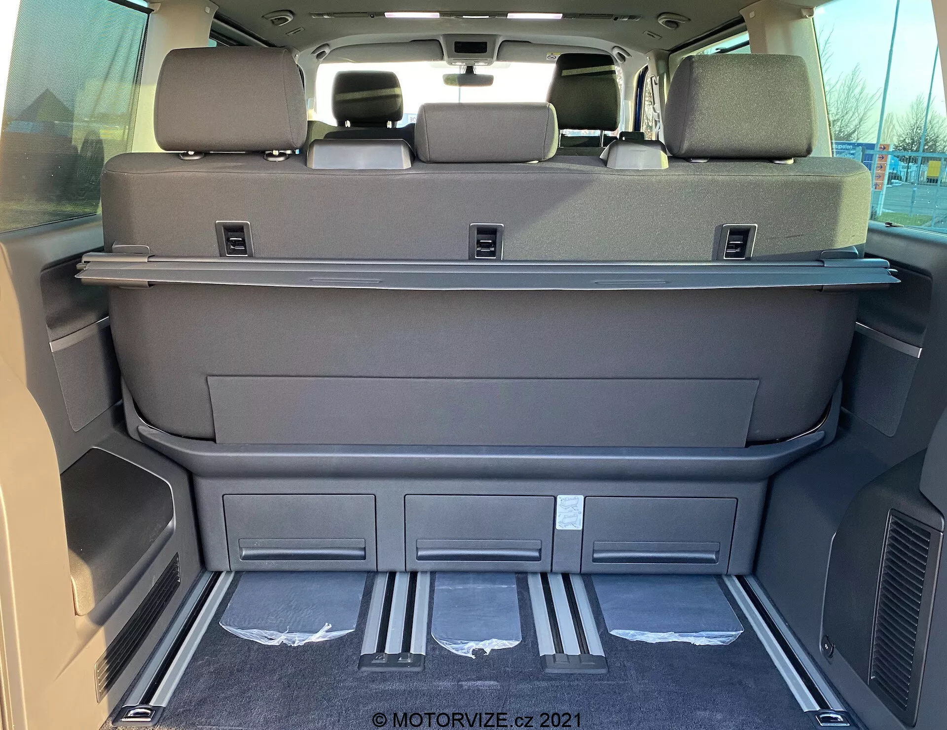 Vista interna dello spazio di carico di un 2019 VW Nutzfahrzeuge Transporter T6.1 Multivan dall'area posteriore, che mostra uno spazio ampio con i sedili della terza fila ripiegati, colore grigio scuro o nero, mensola copribagagli, poggiatesta sui sedili posteriori, protezioni d'ingresso in metallo, scomparti sagomati, prese d'aria, tappetino pulito e possibili controlli elettronici dei sedili o del portellone.