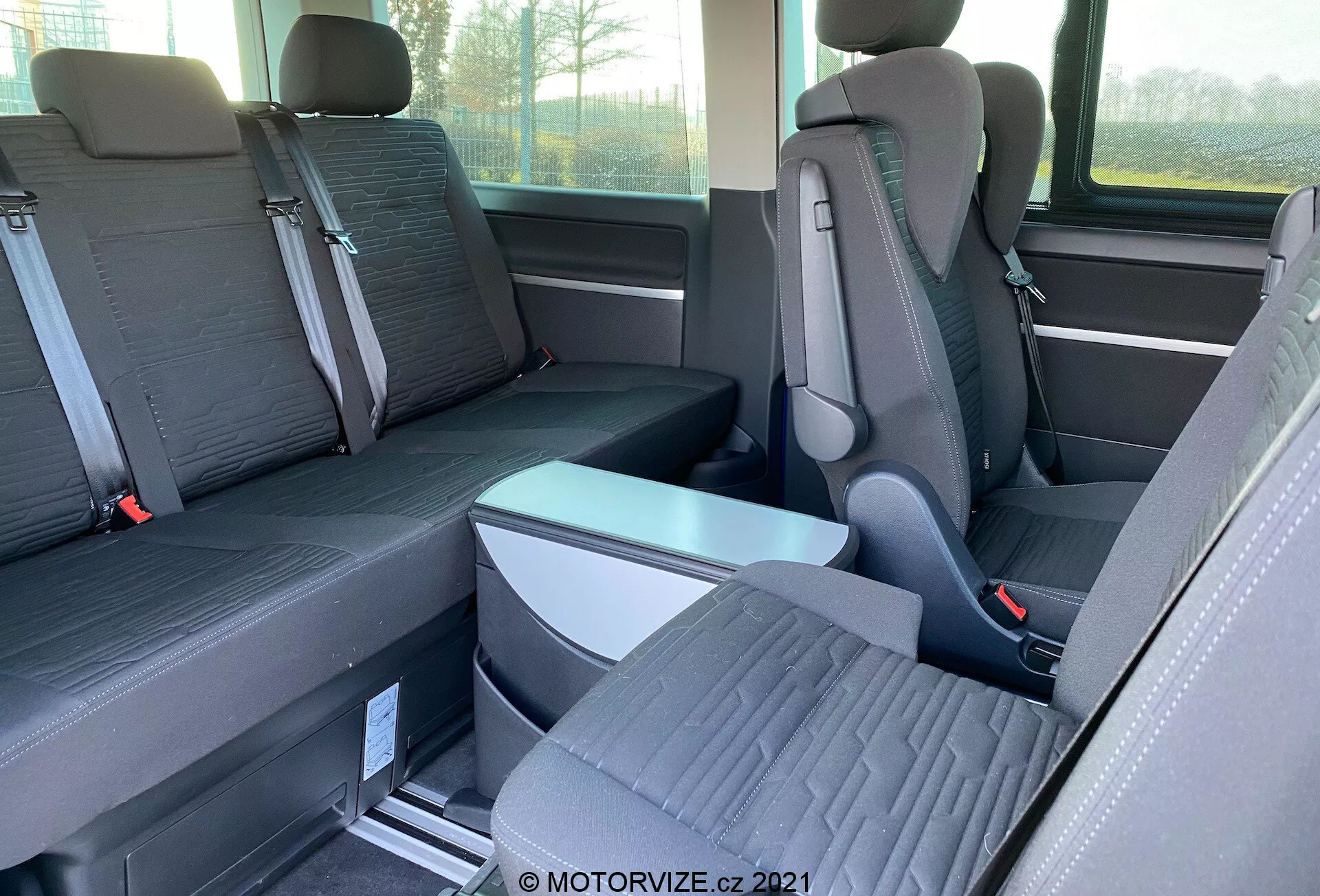 Vista interna del VW-Nutzfahrzeuge Transporter 2019 (T6.1 Multivan) focalizzata sui sedili della seconda e terza fila. Fotografato di lato all'altezza degli occhi, l'immagine mostra sedili individuali con poggiatesta prominenti nella seconda fila e una panca per tre passeggeri nella terza fila. I sedili sono in tessuto bicolore e il pavimento presenta guide metalliche per sedili riconfigurabili. Sono visibili un bracciolo pieghevole e le fibbie delle cinture di sicurezza. Si intravedono anche parzialmente la finestra e lo stoccaggio laterale.