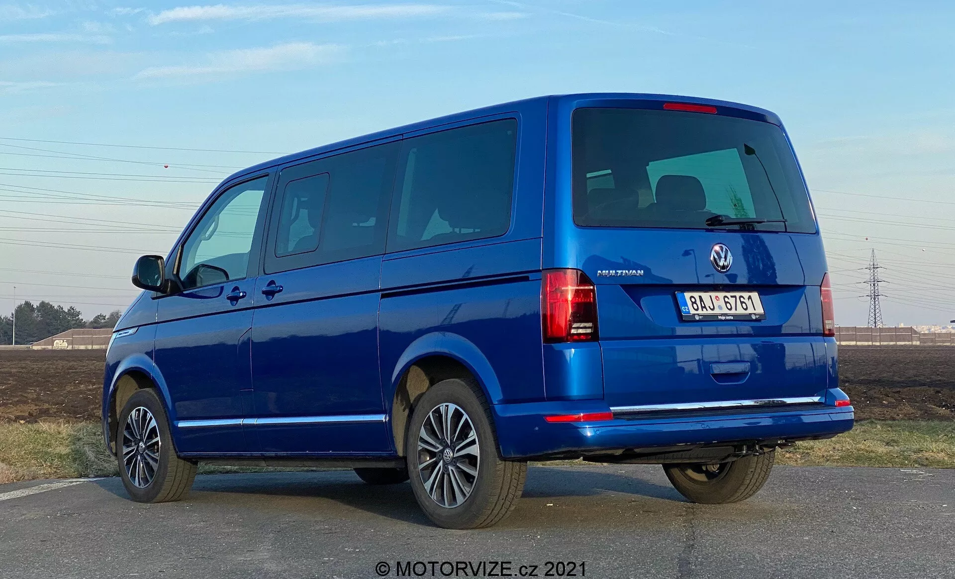 Un furgone blu VW-Nutzfahrzeuge Transporter 2019 (T6-1 Multivan) visto da un angolo posteriore di tre quarti. L'immagine cattura i gruppi ottici posteriori, i finestrini posteriori e il portellone, nonché il profilo laterale con le porte passeggeri e i finestrini. Il furgone occupa gran parte dell'inquadratura a livello degli occhi, rivelando porte posteriori divise e diversi finestrini laterali, indicando un modello passeggeri. I cerchi in lega, i paraurti color carrozzeria e gli specchietti laterali suggeriscono un alto livello di allestimento. L'illuminazione suggerisce che sia di giorno con il cielo sereno e ci sono ombre nitide sotto il veicolo.
