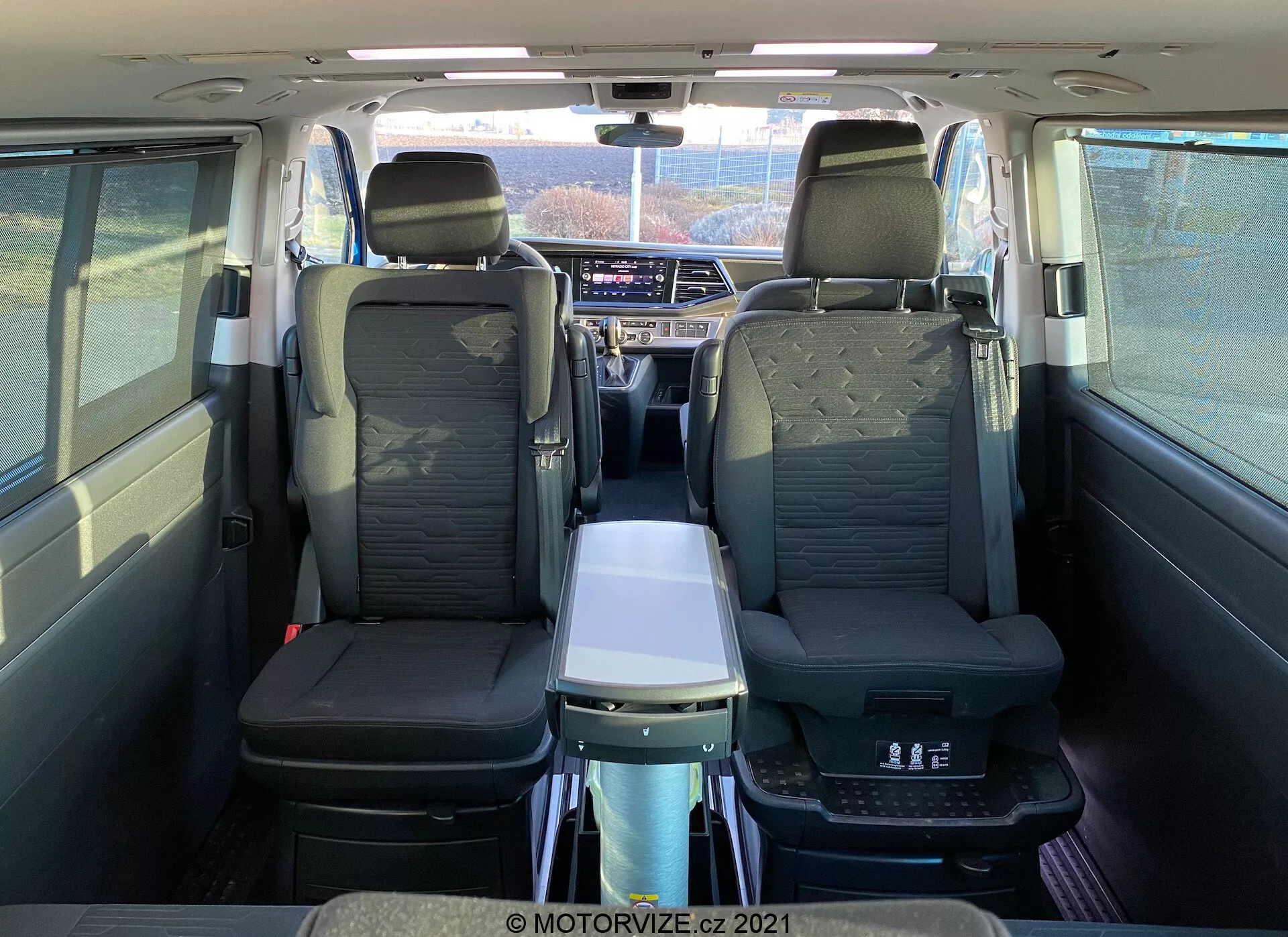 Interno del VW Nutzfahrzeuge Transporter T6-1 Multivan 2019 visto da dietro, che mostra sedili in pelle, braccioli nella seconda fila, tavolino pieghevole, tetto panoramico, bocchette d'aria individuali e avanzato sistema di infotainment; evidenzia il comfort dei passeggeri e l'abitacolo spazioso.