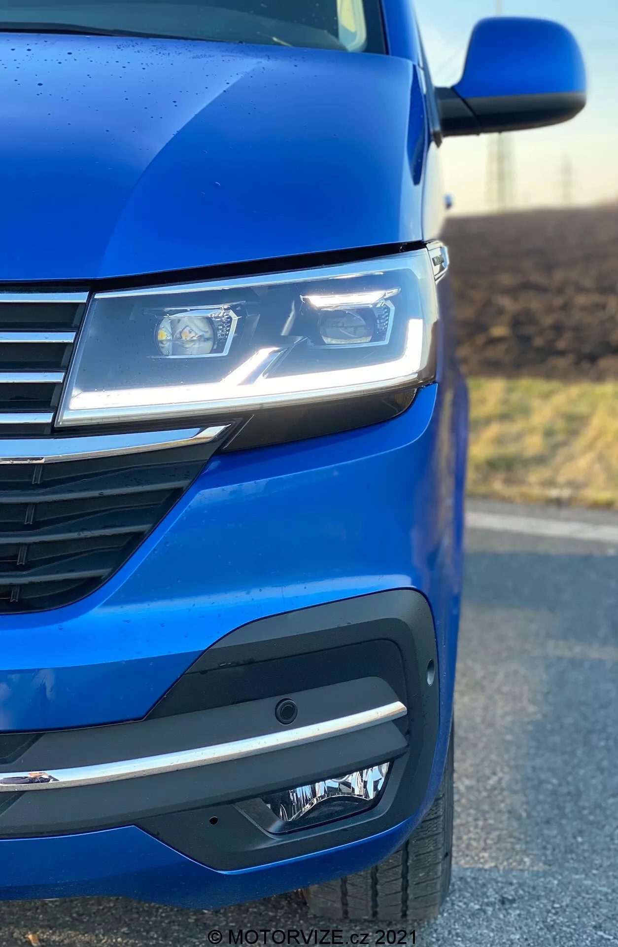 Primo piano del lato anteriore sinistro di un VW Nutzfahrzeuge Transporter 2019 (T6.1 Multivan) che evidenzia la vernice blu metallizzata, i fari LED avanzati, il design moderno della griglia con linee orizzontali e il sofisticato paraurti anteriore con fari fendinebbia o unità sensori. L'angolo della foto enfatizza l'aerodinamica e i dettagli di design dell'automobile.