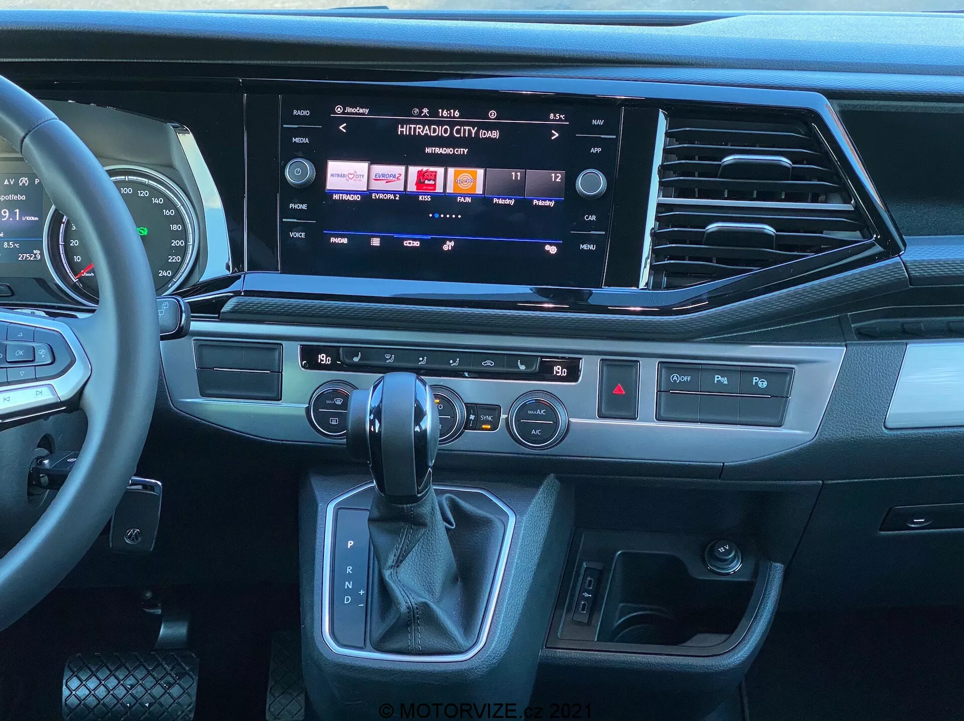Vista interna del VW Nutzfahrzeuge Transporter 2019 T6.1 Multivan dalla prospettiva del sedile passeggero, che mostra il moderno cruscotto e la consolle centrale. Volante multifunzione rivestito in pelle con comandi multimediali, ampio sistema di infotainment a schermo tattile che mostra opzioni multimediali e di connettività, eleganti bocchette dell'aria, pulsanti tattili per funzioni rapide, pannello di controllo del clima a doppia zona, area ergonomica della leva del cambio automatico e pulsante di avviamento/arresto senza chiave. Il design presenta una combinazione di plastiche scure, finiture che sembrano metallo spazzolato e materiali simili al cuoio, evidenziando un ambiente contemporaneo e premium.