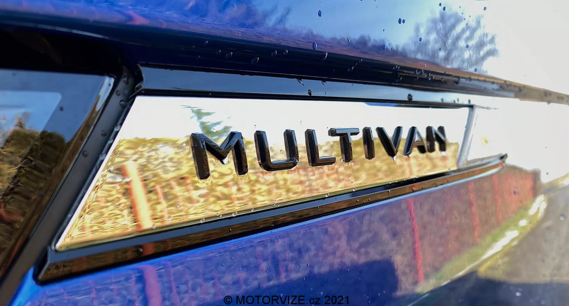Vista ravvicinata di una parte del VW Nutzfahrzeuge Transporter 2019 (T6-1 Multivan) focalizzata sul badge 'MULTIVAN'. L'immagine è scattata da un basso angolo posteriore, guardando leggermente verso l'alto dove la finestra posteriore si incontra con il bordo del tetto. Leggeri riflessi sul vetro della finestra suggeriscono un ambiente esterno. Questa immagine strettamente ritagliata enfatizza l'emblema di designazione del modello del produttore.