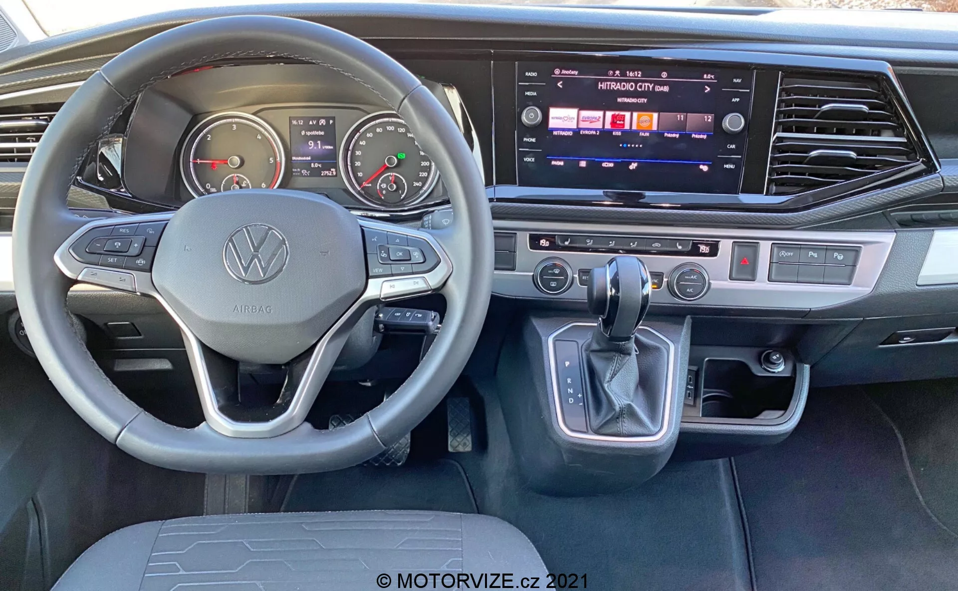 Vista interna del VW Nutzfahrzeuge Transporter 2019 T6.1 Multivan dalla prospettiva del sedile del conducente. Presenta un volante con logo Volkswagen e pulsanti multifunzione, strumenti analogici, display digitale, console centrale con sistema di infotainment touchscreen attivo, climatizzatore bizona, leva del cambio con pulsante di parcheggio elettronico e accenti moderni in metallo spazzolato.