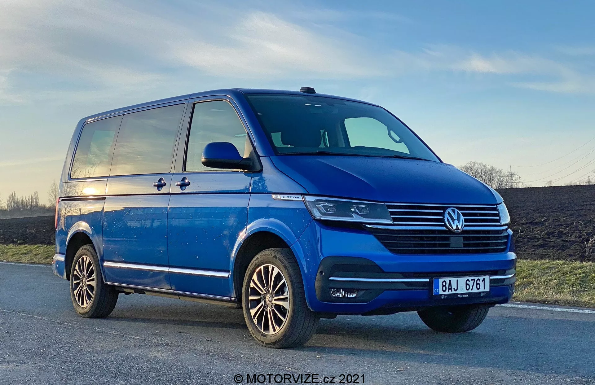 Vista in basso di un Volkswagen T6-1 Multivan blu del 2019, che mostra il quadrante anteriore sinistro da una prospettiva a tre quarti. Il furgone è parcheggiato su una strada leggermente inclinata, con dettagli cromati prominenti sulla griglia, un emblema VW centrale e fari moderni che possono utilizzare la tecnologia LED. I fari fendinebbia sono integrati nel paraurti, che ha anche prese d'aria. Il profilo laterale rivela una porta scorrevole, una linea di carattere che si estende dal parafango anteriore a quello posteriore e specchietti laterali sostanziali con indicatori di direzione. Sono visibili anche i cerchi in lega con un design a più raggi e il battistrada degli pneumatici ben mantenuto. Nel fondo, il cielo mostra una leggera sfumatura di tramonto.