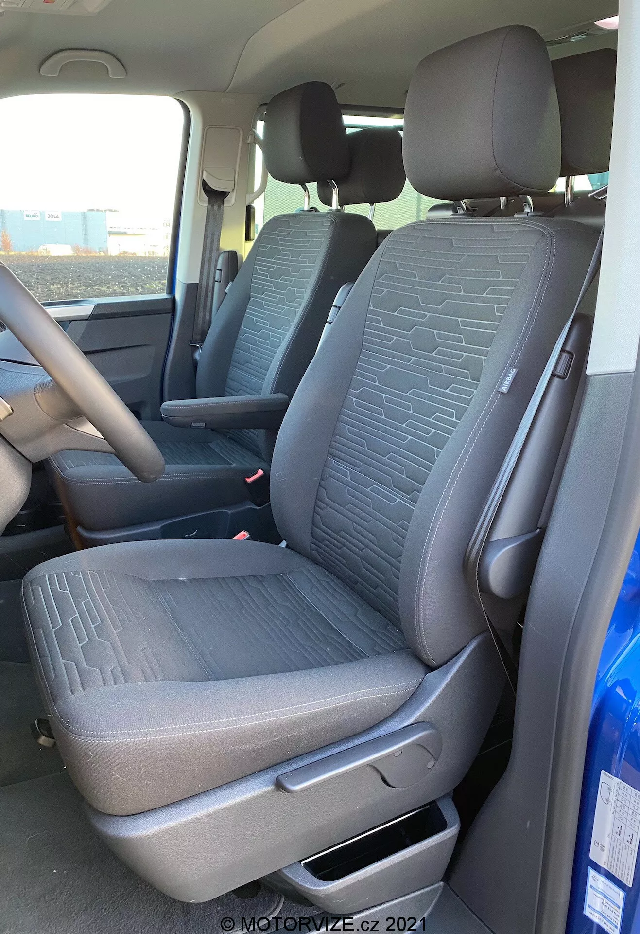Interni del VW Nutzfahrzeuge Transporter 2019 T6.1 Multivan, che mostrano il sedile anteriore del passeggero e parte del sedile del conducente da una prospettiva esterna sul lato del passeggero. La ripresa, effettuata da un angolo leggermente basso, mette in risalto il tessuto ruvido con motivi geometrici sui sedili, la leva di regolazione manuale, le cinture di sicurezza e parte della console centrale e del volante. La luce naturale illumina l'interno del veicolo.