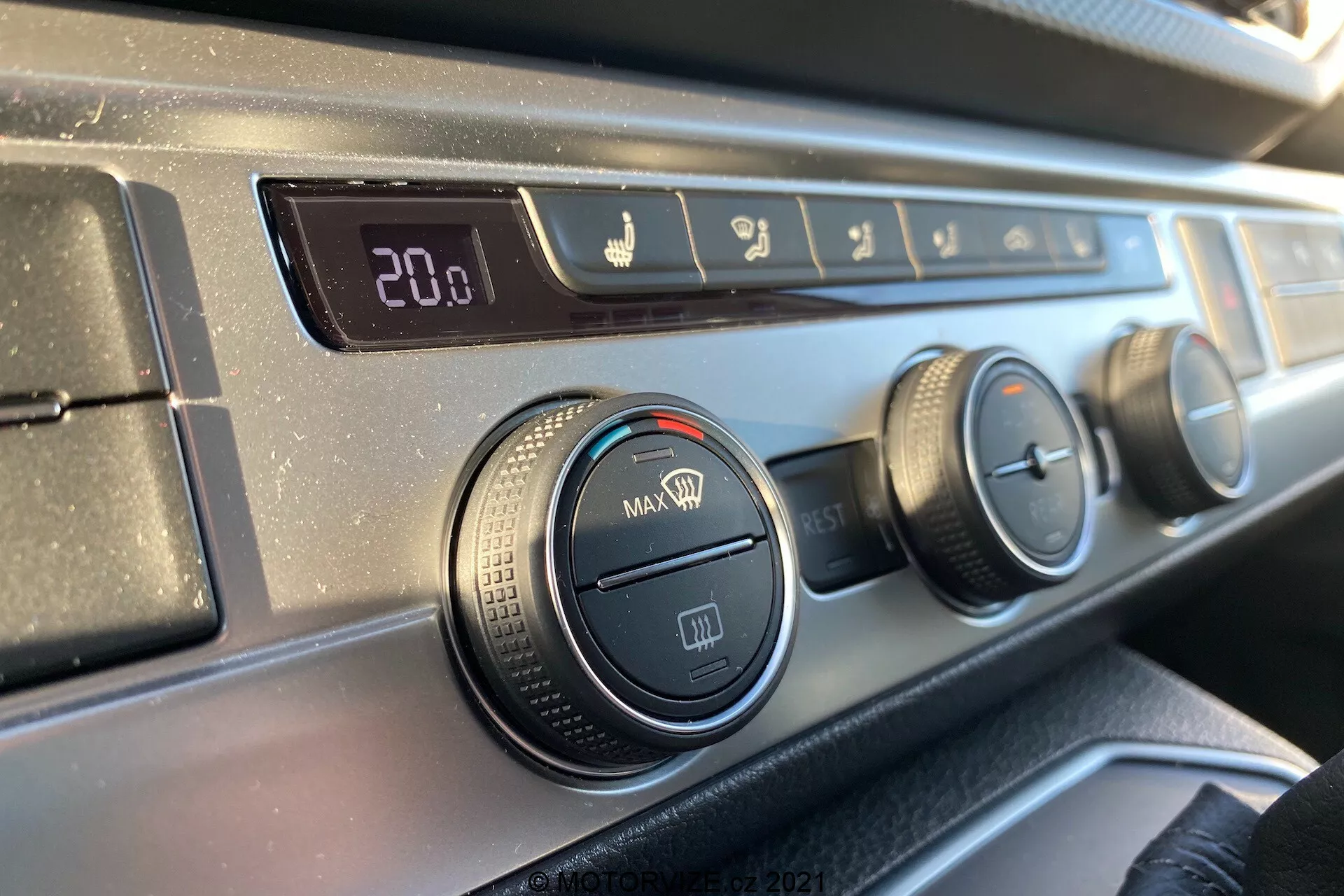 Vista ravvicinata del pannello di controllo del clima sul cruscotto del VW Transporter T6.1 Multivan 2019 dalla prospettiva di un occupante del sedile anteriore, che mostra un display digitale della temperatura impostato su 20.0 gradi Celsius, controlli della direzione del flusso d'aria, tre manopole rotanti per impostazioni di aria condizionata e ventola e un pulsante di ricircolo, tutto con bordi in plastica nera testurizzata e una finitura liscia e scura del cruscotto.