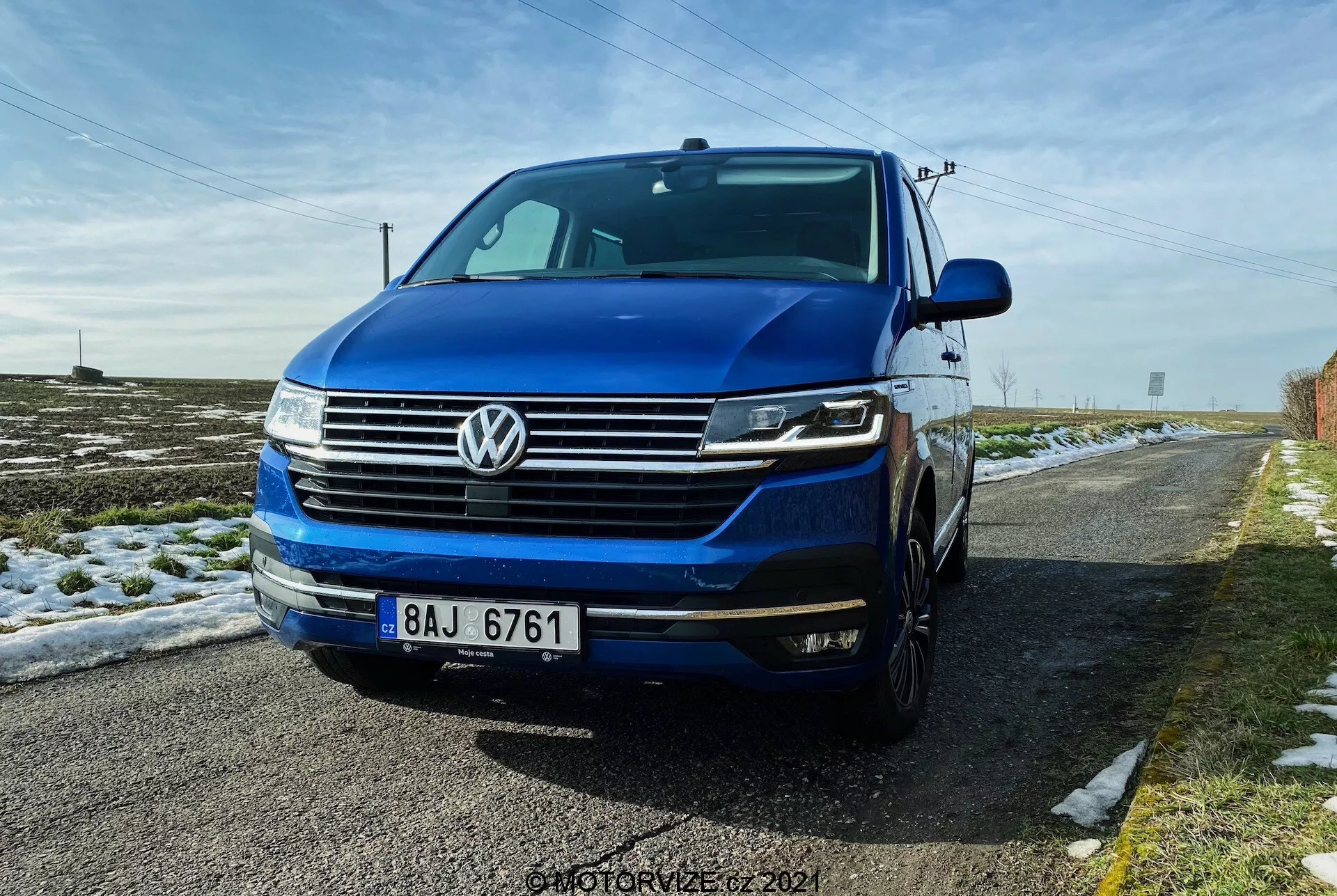 Una vista a tre quarti anteriore bassa del Volkswagen T6.1 Multivan 2019, con la griglia anteriore con linee orizzontali, il distintivo emblema VW centrato sulla griglia, i fari con luci diurne a LED e probabilmente fari bi-xenon o LED, e il paraurti anteriore con fendinebbia e prese d'aria. L'immagine mostra parte del lato sinistro, compresa la porta del conducente e il parafango, con il cofano e il parabrezza visibili. L'auto è di un blu intenso e parcheggiata su una strada rurale con neve sul terreno e un cielo azzurro limpido sopra.