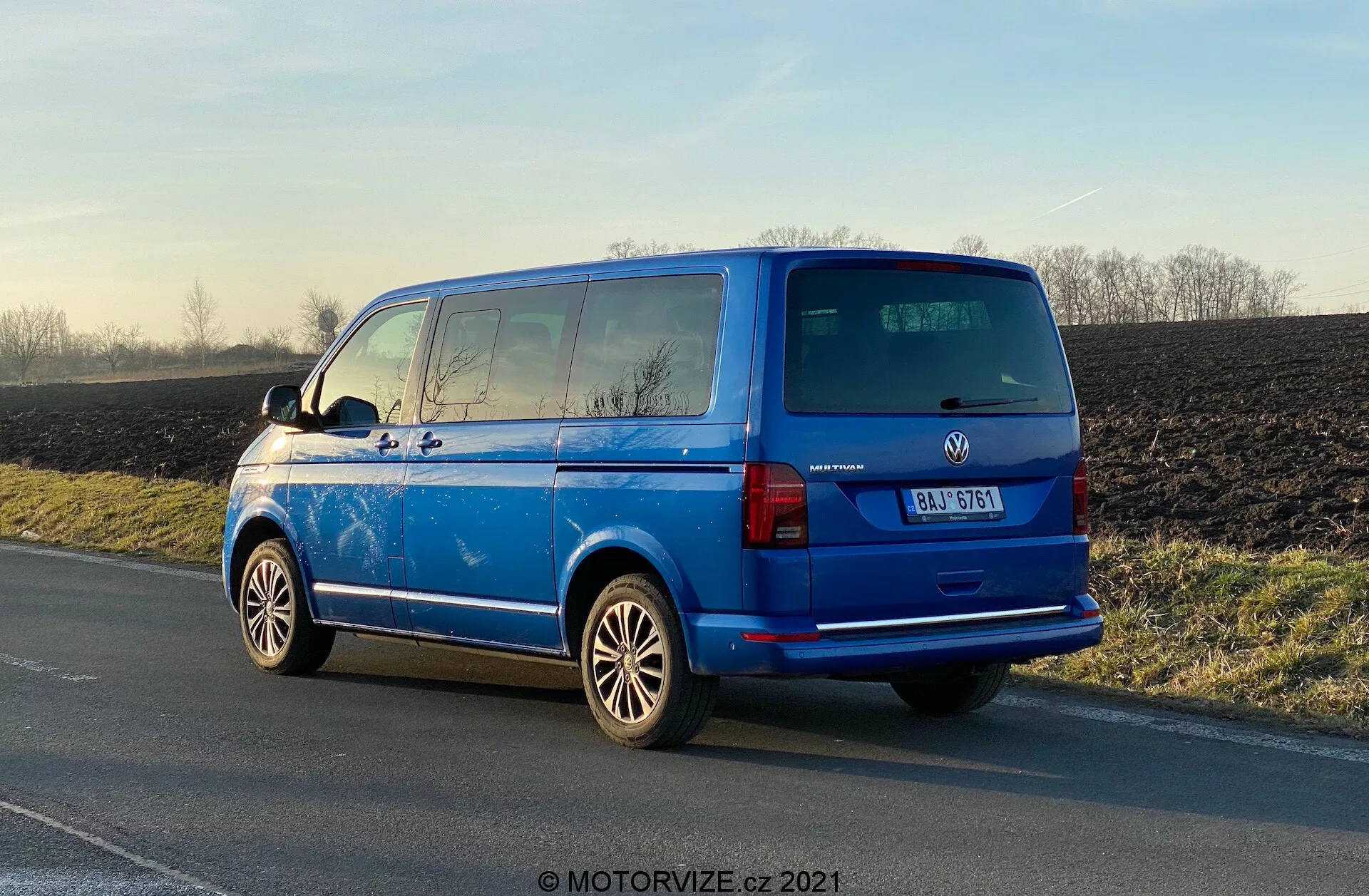 Volkswagen Transporter 2019 (T6-1 Multivan) blu vibrante parcheggiato sul ciglio della strada, vista posteriore tre quarti che mostra luci posteriori, finestrino posteriore, portellone, badge del produttore, dettagli del modello del veicolo; cerchi in lega prominenti, specchietti laterali esterni, linee del sbrinatore posteriore, finestrini oscurati in seconda fila, antenna sul tetto; parcheggiato alla luce naturale con riflessi che evidenziano la superficie lucida, sfondo rurale.
