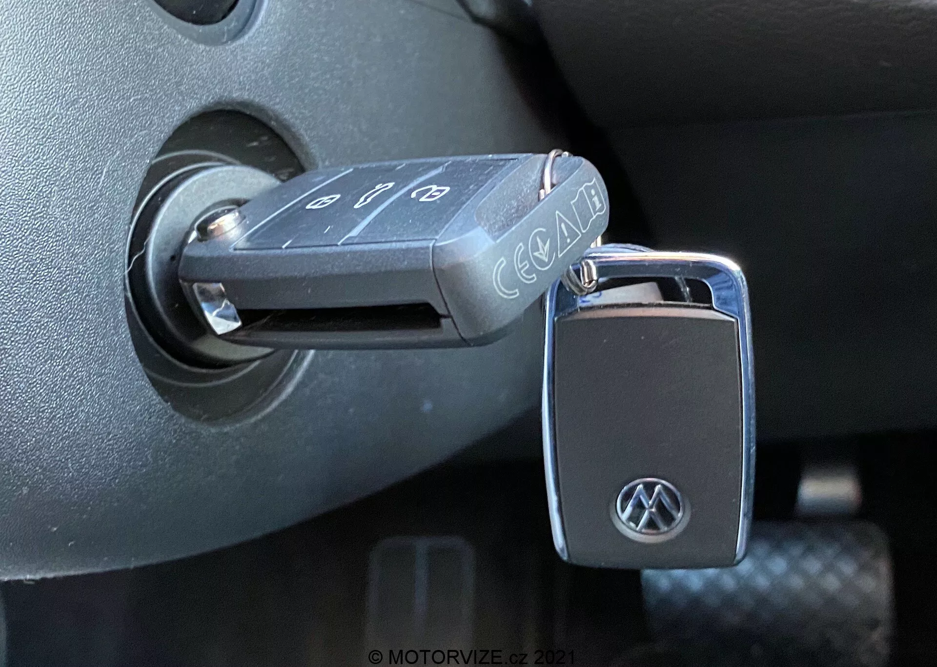 Primo piano di una chiave pieghevole Volkswagen inserita nell'interruttore di accensione di un VW Nutzfahrzeuge T6-1 Multivan del 2019. La chiave, con pulsanti per bloccare, sbloccare e allarme panico, mostra un logo VW sul portachiavi. L'interruttore di accensione si trova nell'ambiente plastico scuro della colonna dello sterzo, indicando che l'accensione è in posizione 'spenta'. La foto, scattata da un'angolazione laterale con luce naturale, mette in evidenza le trame e le finiture dettagliate degli interni del veicolo.