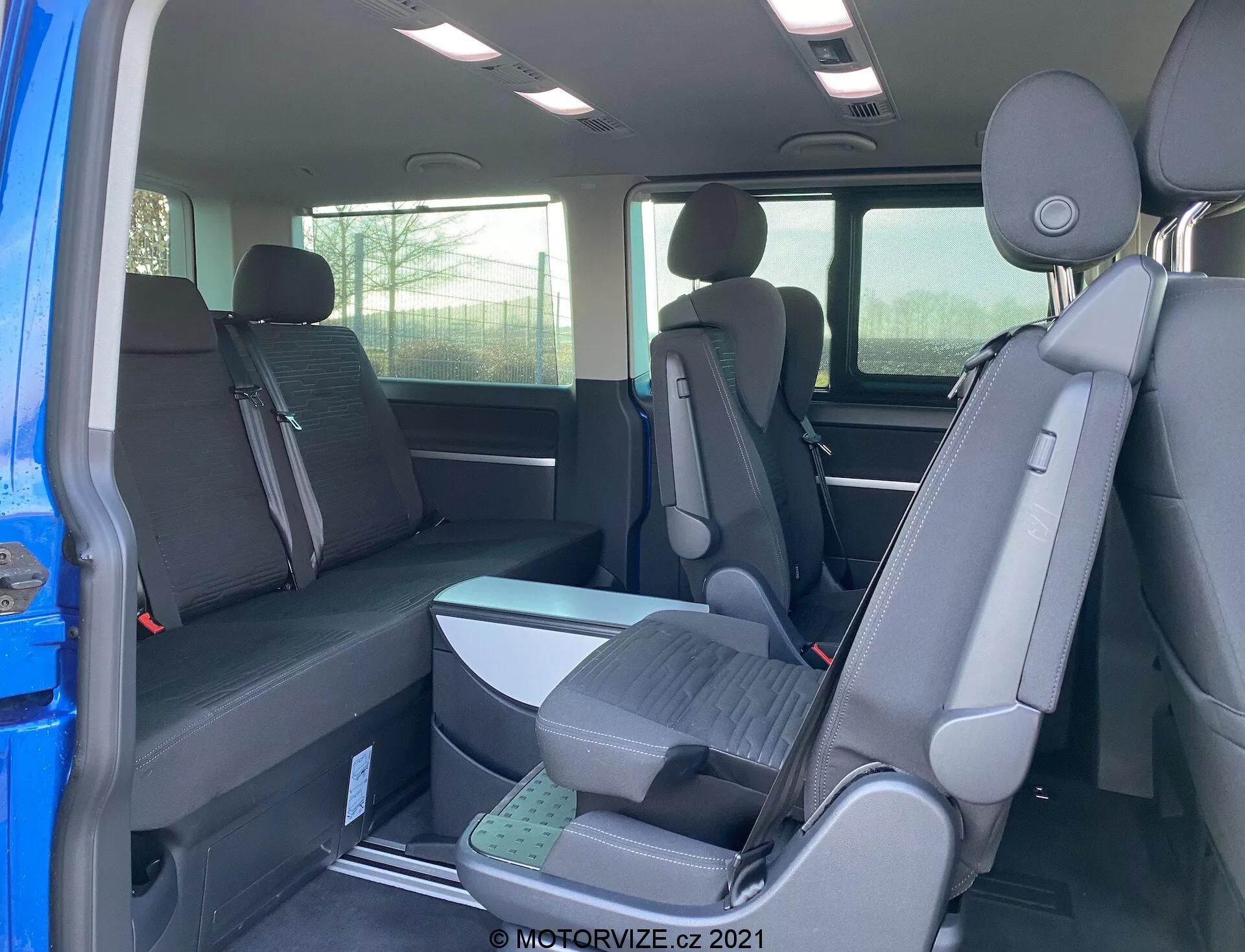 Interno del VW Transporter T6.1 Multivan 2019 che mostra i sedili della seconda e terza fila dalla prospettiva del lato passeggero posteriore, con configurazione dei sedili 2-3-2 e rivestimento in tessuto scuro. Vista parziale dell'esterno attraverso la porta aperta, poggiatesta su tutti i sedili e sedili della terza fila rialzati per una migliore visibilità.
