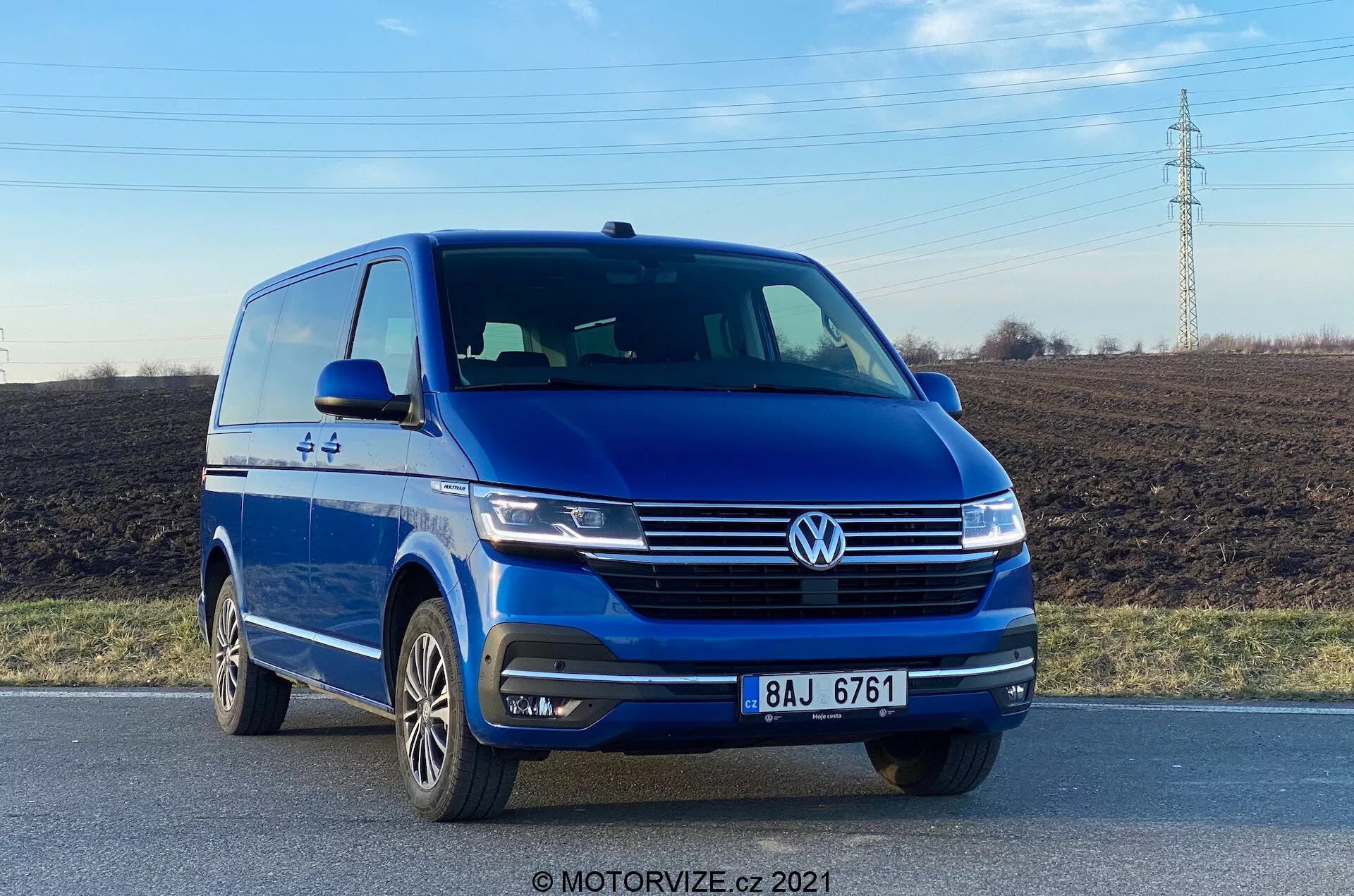 L'immagine mostra un furgone blu Volkswagen VW-Nutzfahrzeuge Transporter 2019 (T6-1 Multivan) catturato da un'angolazione frontale bassa, evidenziando il quarto anteriore sinistro con una vista chiara della fascia frontale e del profilo del lato guida. Il veicolo è su una strada, mettendo in risalto elementi come il distintivo Volkswagen prominente, i fari eleganti, le linee scolpite sul cofano, i fendinebbia integrati nel paraurti e le ruote in lega multi-razza. La fotografia, scattata alla luce del giorno, enfatizza lo stile esterno del furgone e le tecnologie di assistenza alla guida, come i sensori di parcheggio.