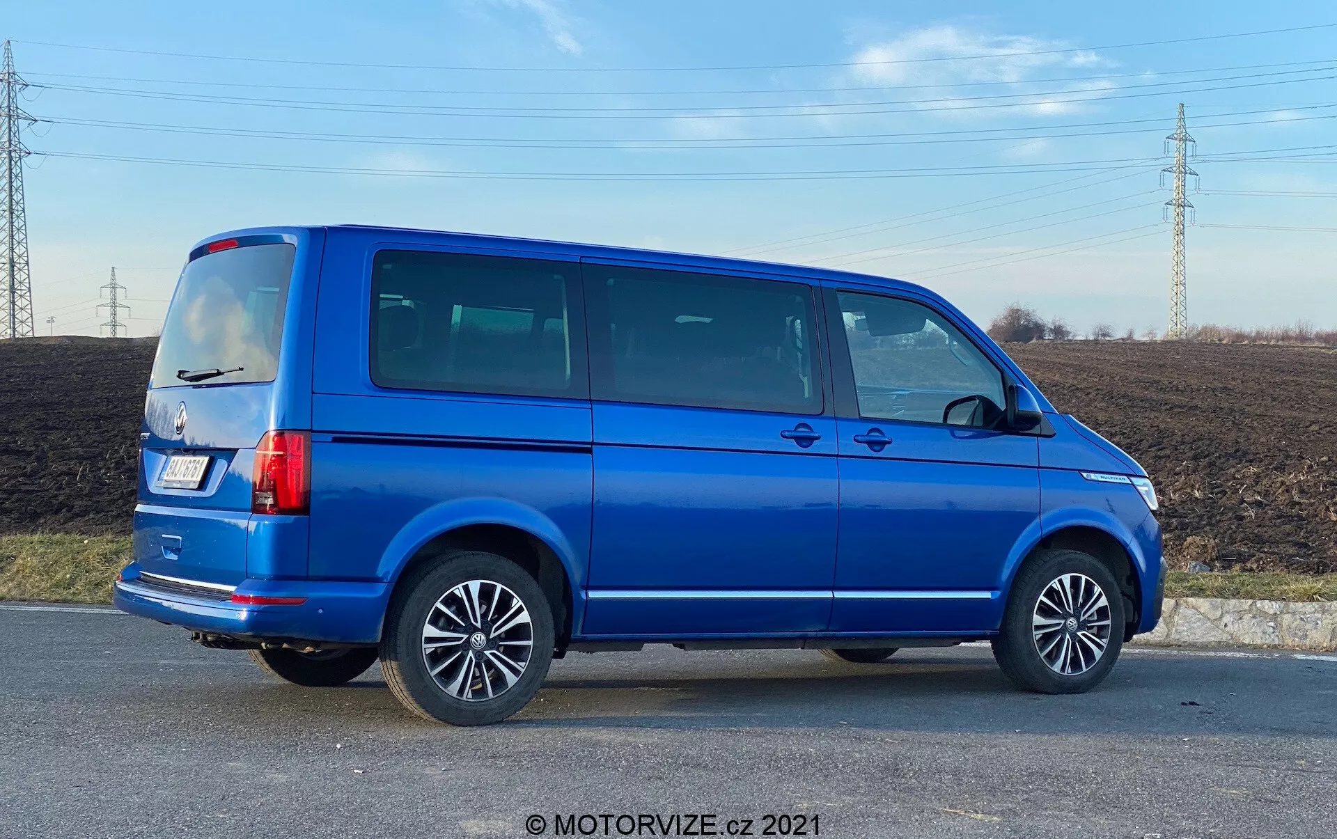 Vista completa del profilo laterale di un Volkswagen Nutzfahrzeuge Transporter blu del 2019 (T6.1 Multivan) con cerchi in lega, linee visibili delle porte, finestrini laterali oscurati, silhouette squadrata, luci posteriori verticali, antenna sul tetto e vernice metallizzata blu brillante. L'immagine è scattata da un angolo che fornisce prospettiva sia sul lato che sulla parte posteriore del veicolo, situato in una zona rurale con un campo e linee elettriche sullo sfondo, in luce diurna.