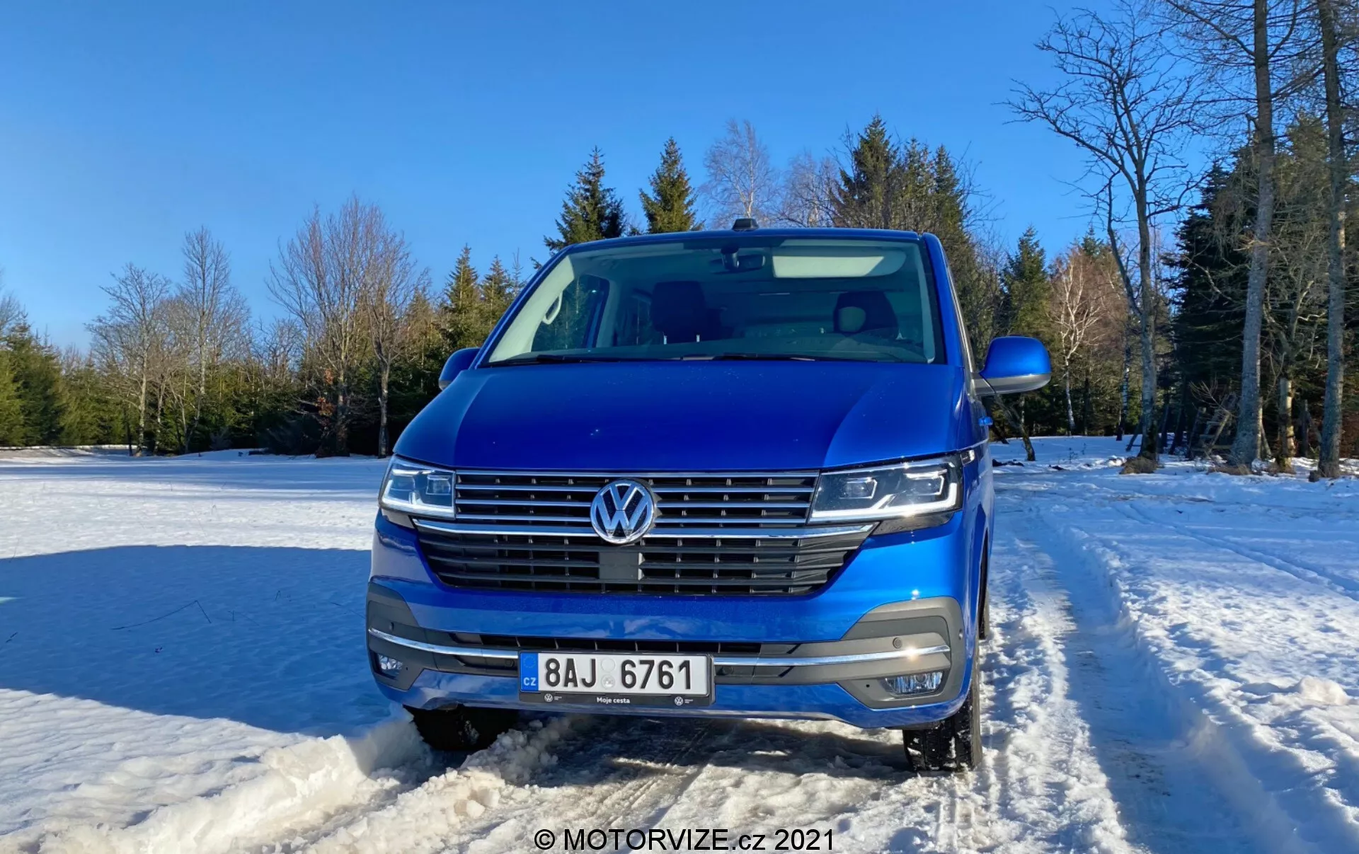 Scatto frontale di un Volkswagen Transporter 2019 (T6-1 Multivan) blu, che mostra la griglia, i fari e l'area del cofano. Posizionato all'aperto su terreno coperto di neve con cielo sereno e alberi sullo sfondo. Le caratteristiche dell'auto includono l'emblema VW, griglia orizzontale con rifiniture cromate, fari LED, telecamera frontale, sensori di parcheggio, barre sul tetto argentate e specchietto laterale parzialmente visibile. Design contemporaneo con parte anteriore aerodinamica, fotografato di giorno con luce solare soffusa dal lato destro.
