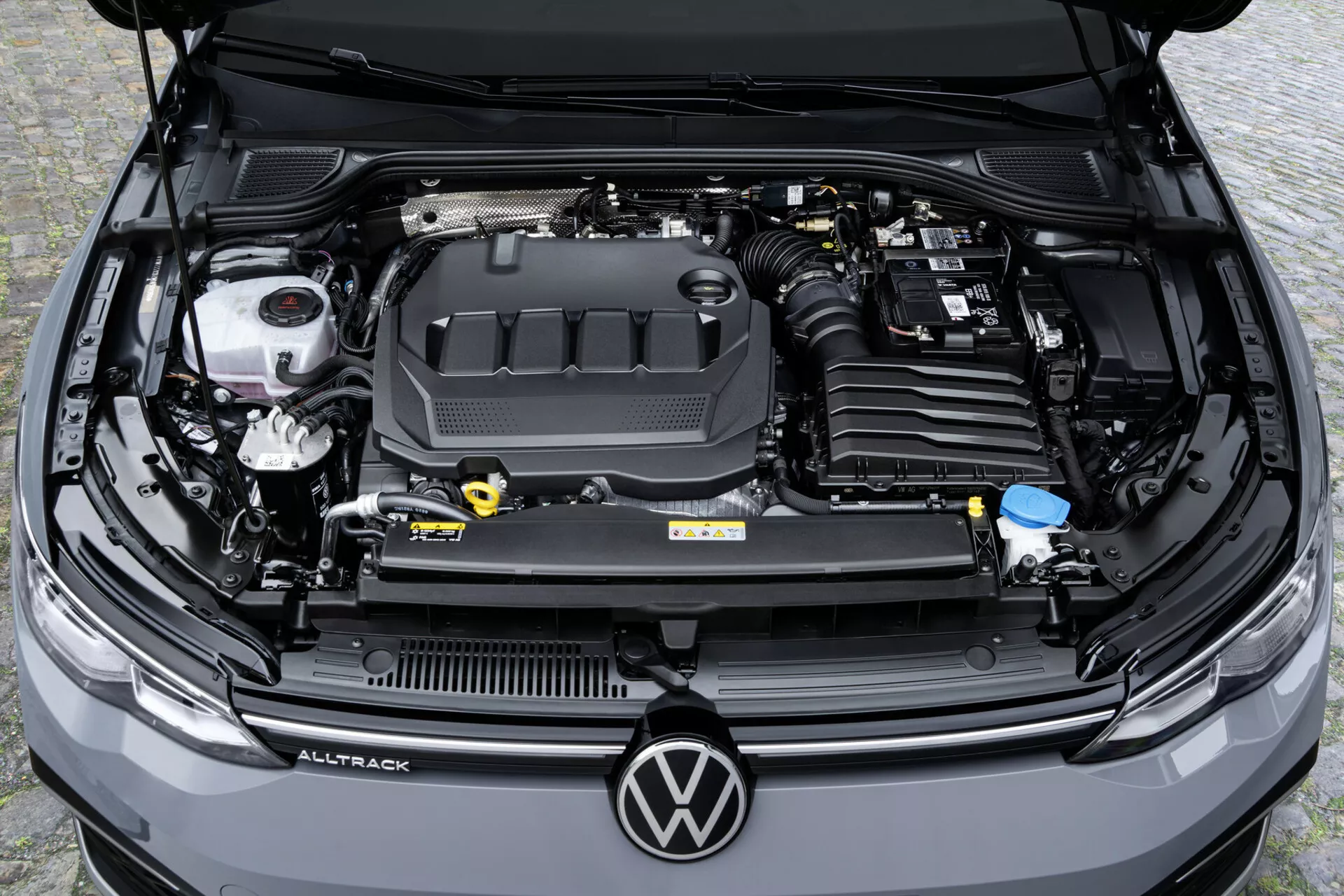 Vista dall'alto del vano motore di una Volkswagen Golf Alltrack 2017 con evidenza del coperchio del motore con logo, serbatoi dei fluidi e batteria dell'auto