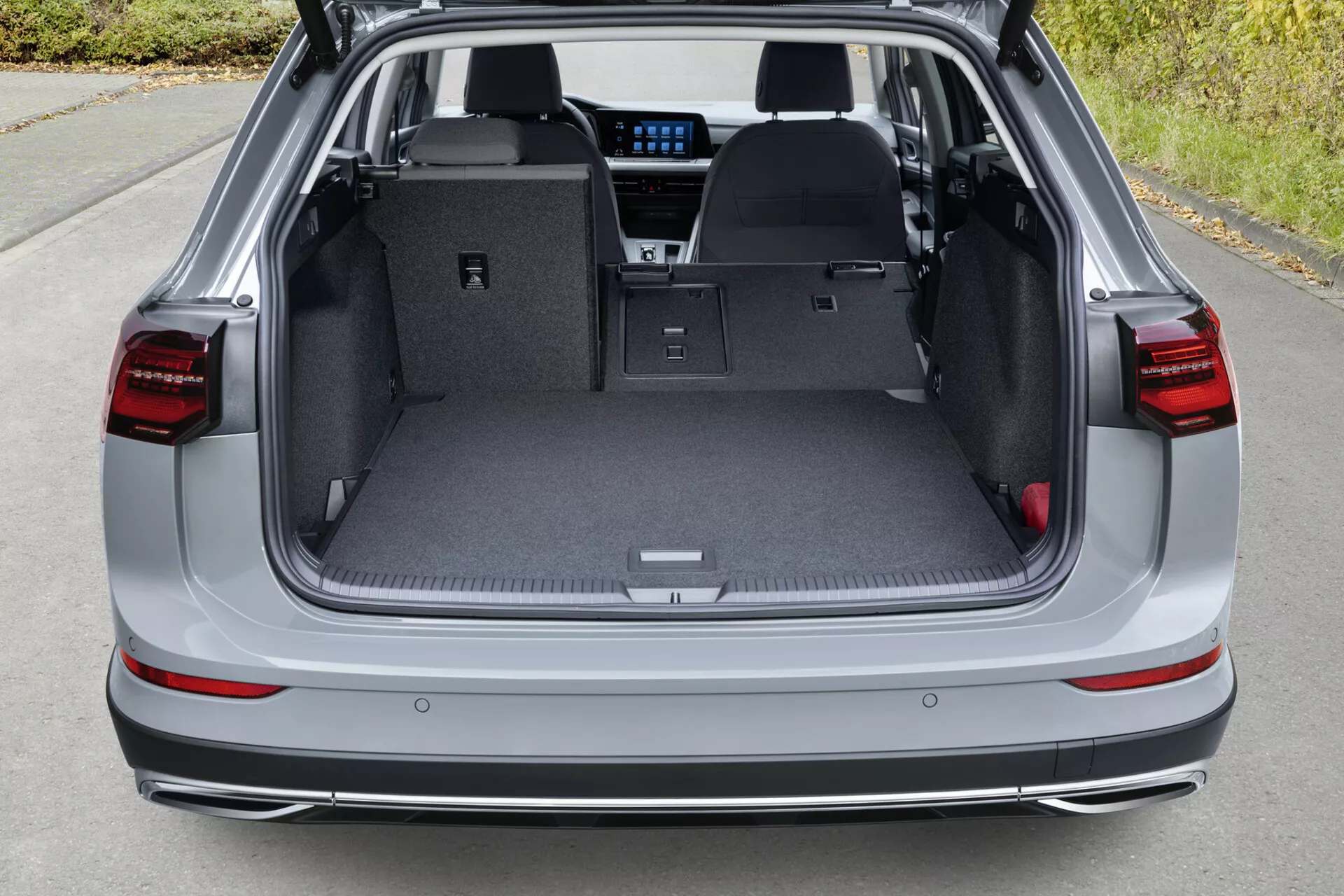 Vista dall'alto dello spazio di carico posteriore di una VW Golf Alltrack 2017 con il portellone aperto, che mostra il piano di carico piatto con i sedili posteriori abbassati, fanali posteriori LED, sensori di parcheggio sul paraurti posteriore e accenni di tecnologia di infotainment moderna attraverso la parte posteriore.