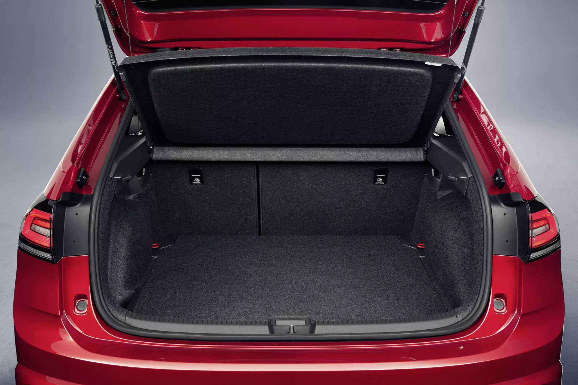 Vista dettagliata dell'esterno rosso e dell'ampio interno del bagagliaio della VW Taigo 2021 con rivestimento in moquette resistente, sedili posteriori abbattibili con cinghie di tiro rosse, copertura del carico retrattile e scomparti di stoccaggio